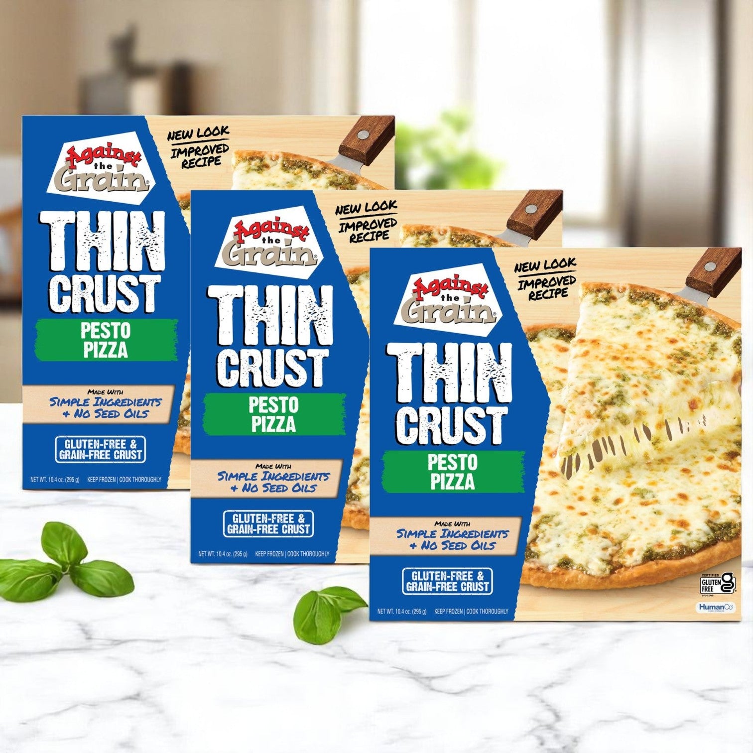 Three packs of Against the Grain Gourmet Pizza, Nut-Free Pesto, Family Size (22.4 oz) are displayed on a kitchen counter with fresh basil leaves in the foreground.