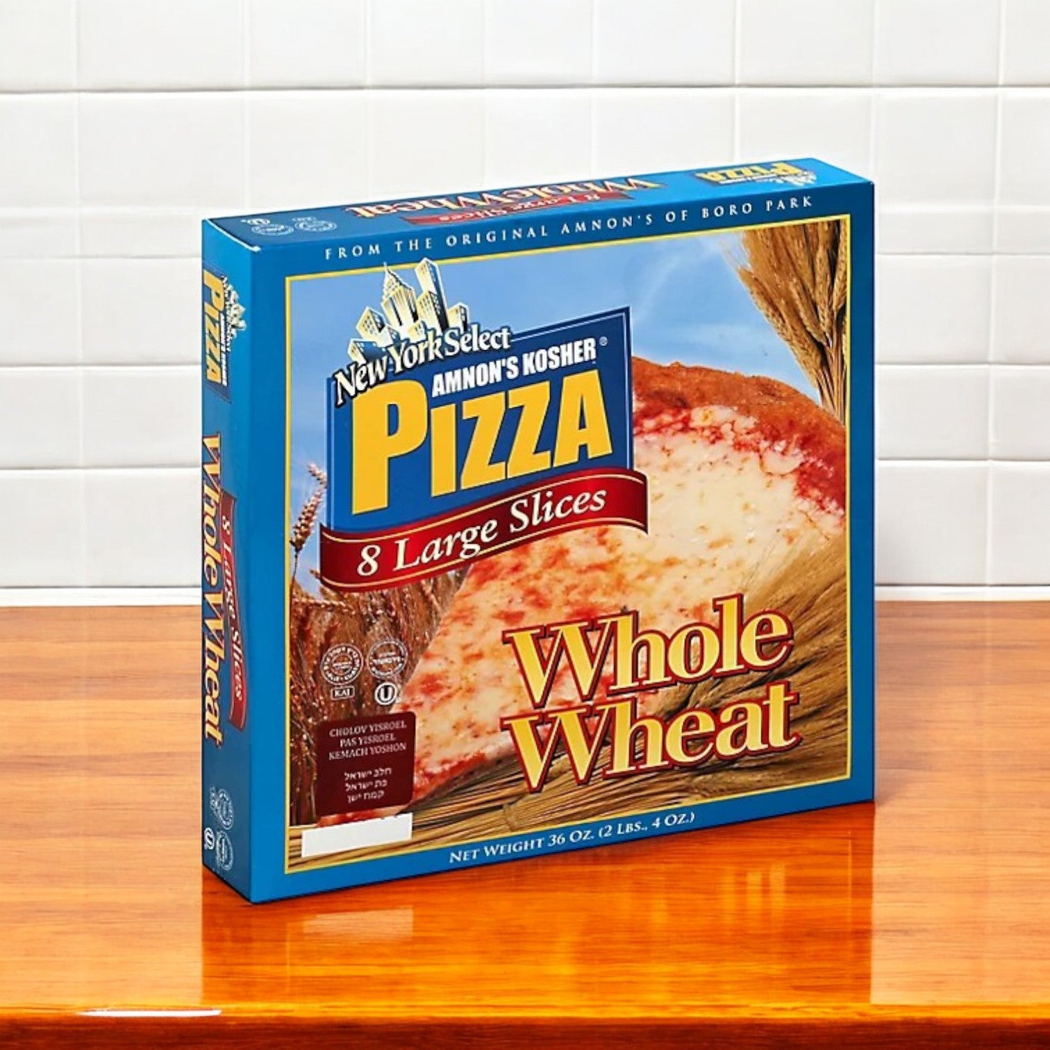 A box from Amnon's Kosher Pizza 3 Pack Bundle, featuring whole wheat, premium mozzarella cheese, and 8 large slices per pizza, sits on a wooden counter against a tiled wall.