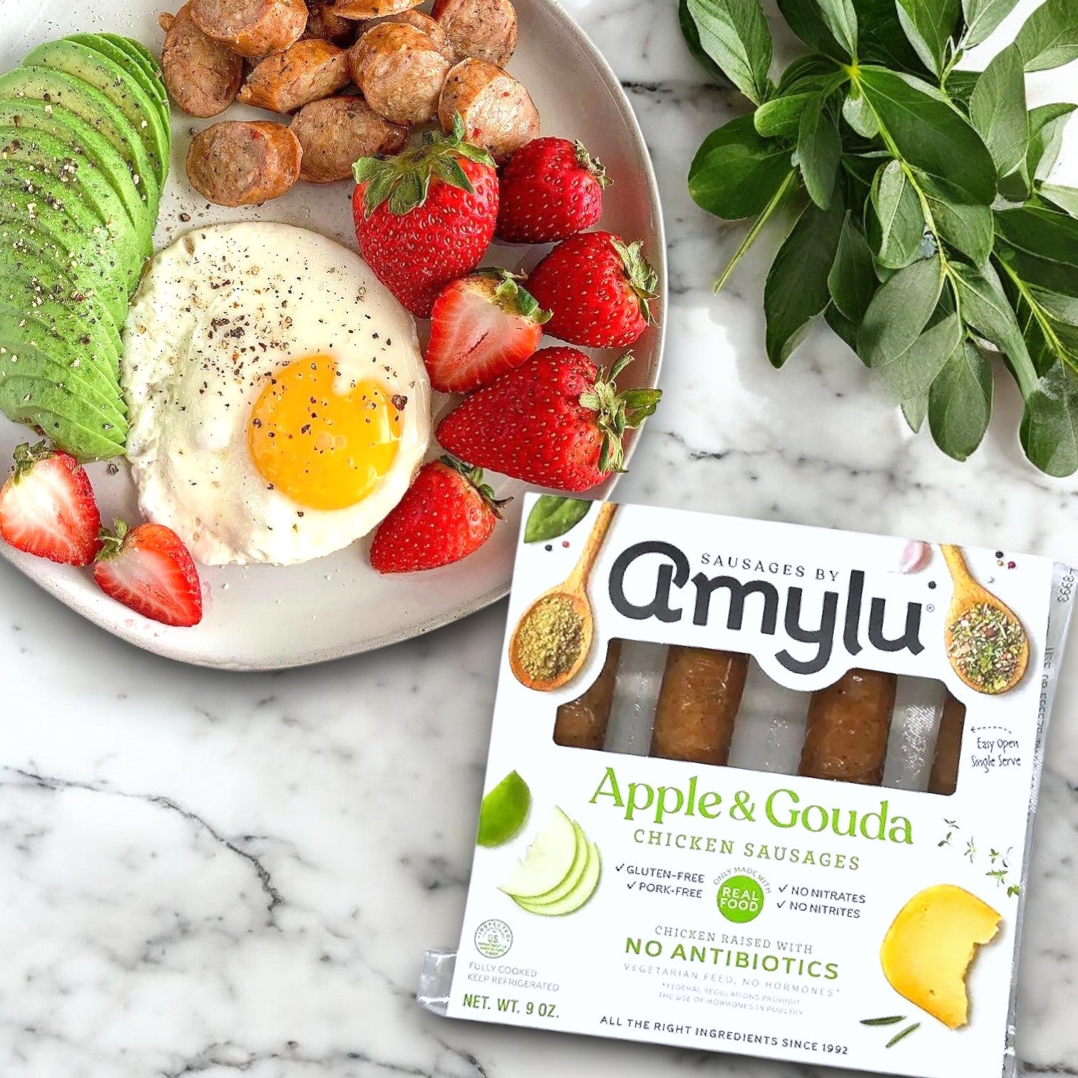 A breakfast plate with sunny-side-up egg, avocado, and strawberries gets a gourmet touch with Amylu Apple and Gouda Chicken Sausage. These fully cooked "Amylu" treats rest enticingly beside their box on the marble surface.