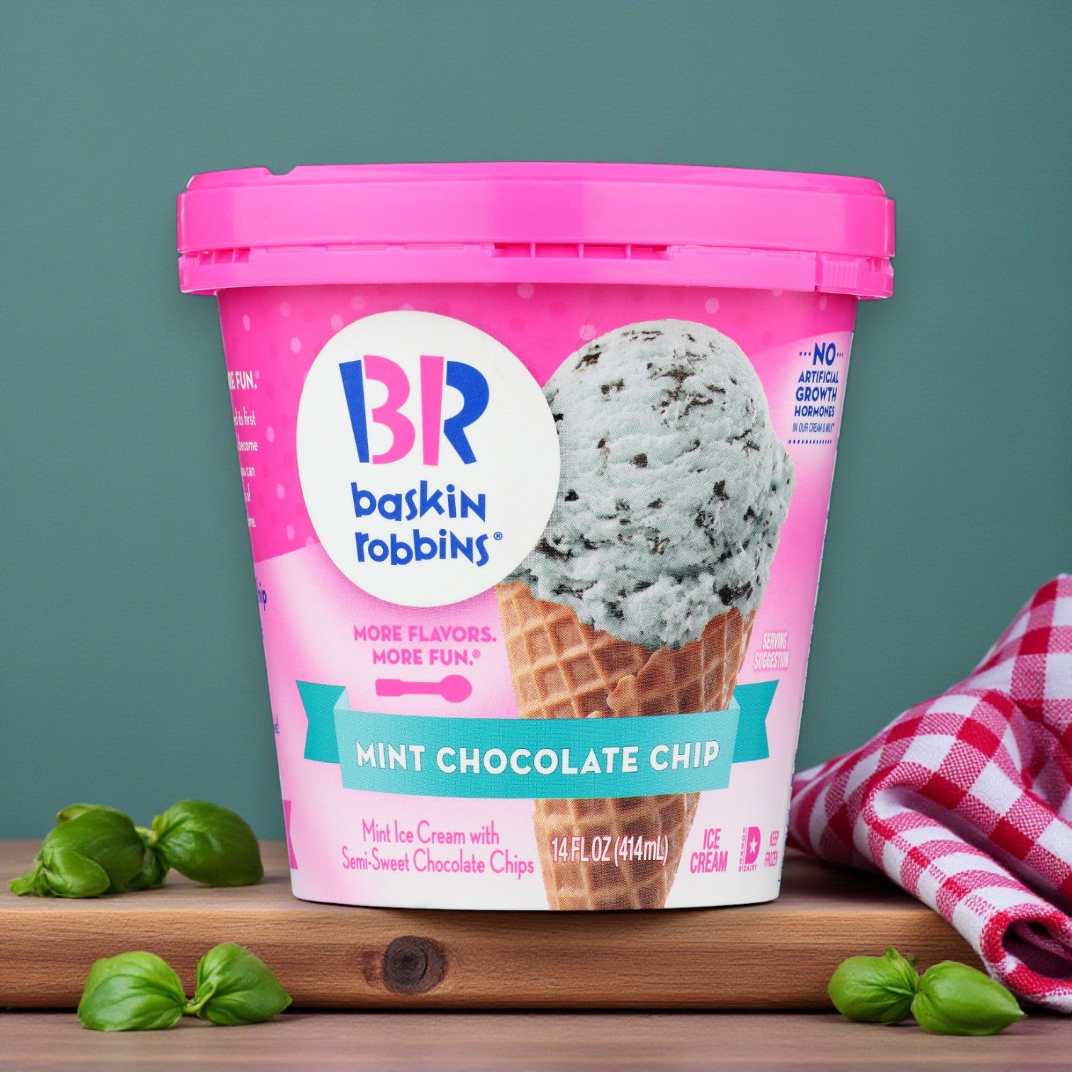 A 14 oz. pint of Baskin Robbins Ice Cream Mint Chocolate Chip sits on a wooden surface next to basil leaves and a red-checkered cloth, highlighting this premium dessert from Baskin Robbins.