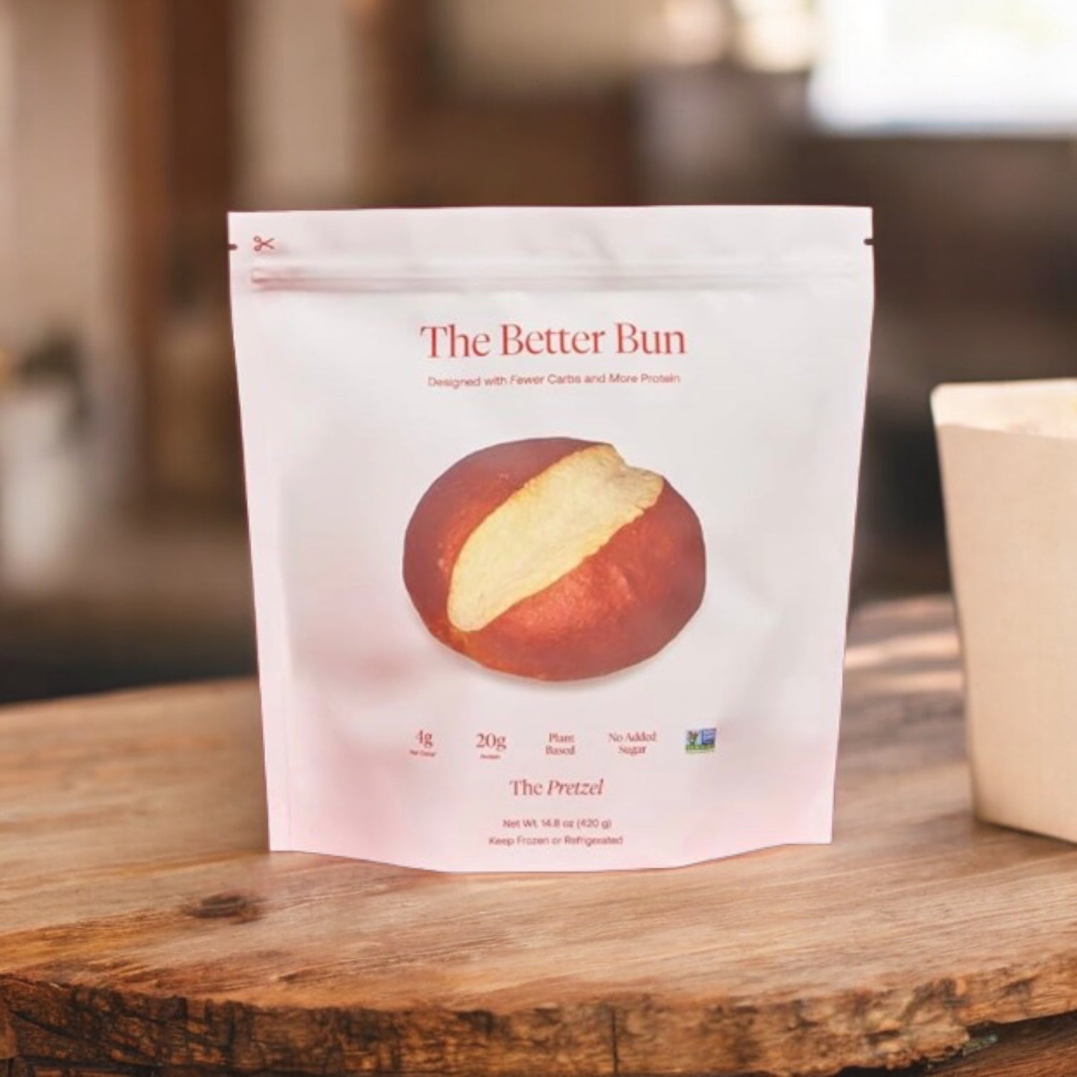A stand-up pouch labeled “BetterBrand, The Better Bun – The Pretzel (1 Bag, 4 Buns)” with a round pretzel-style bun on a wooden surface in soft indoor light. Now features low net carbs for a healthier choice. In stock—ready to ship!.