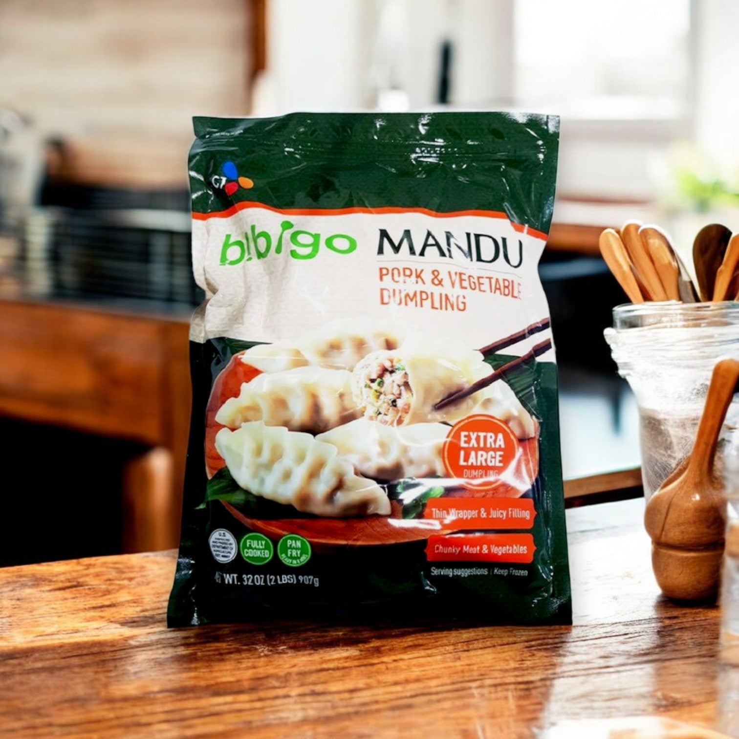 A package of Bibigo Mandu Pork & Vegetable Dumpling | 32 oz, 3 Packs by Bibigo rests on a wooden kitchen counter, highlighting authentic Korean dumplings with kitchen utensils in the background.