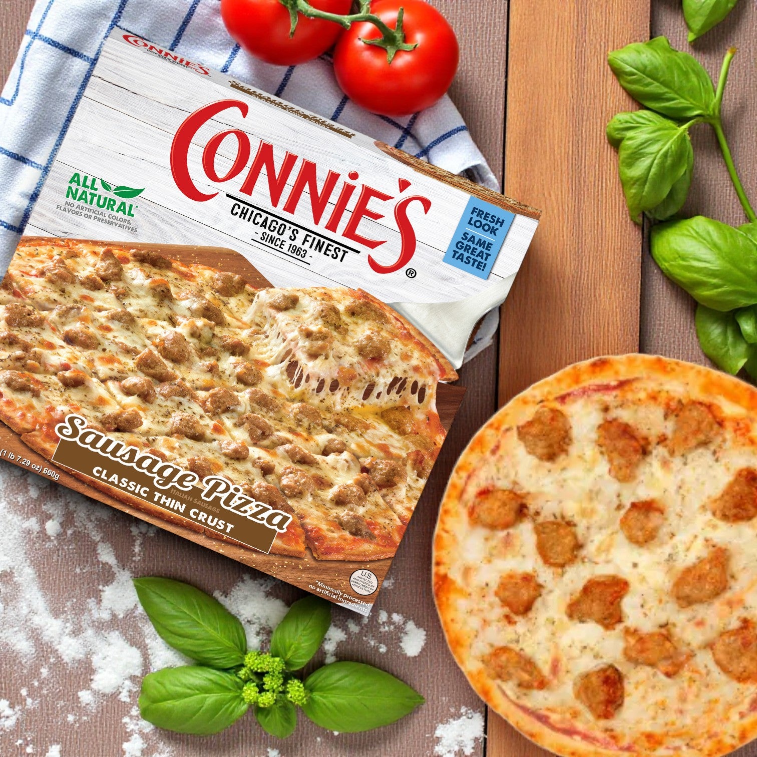 A box of Connie's Pizza Classic Thin Crust Combo – 3 Pack! sits beside a cooked Chicago-style sausage pizza on a wooden surface, surrounded by fresh basil, spinach, and tomatoes.