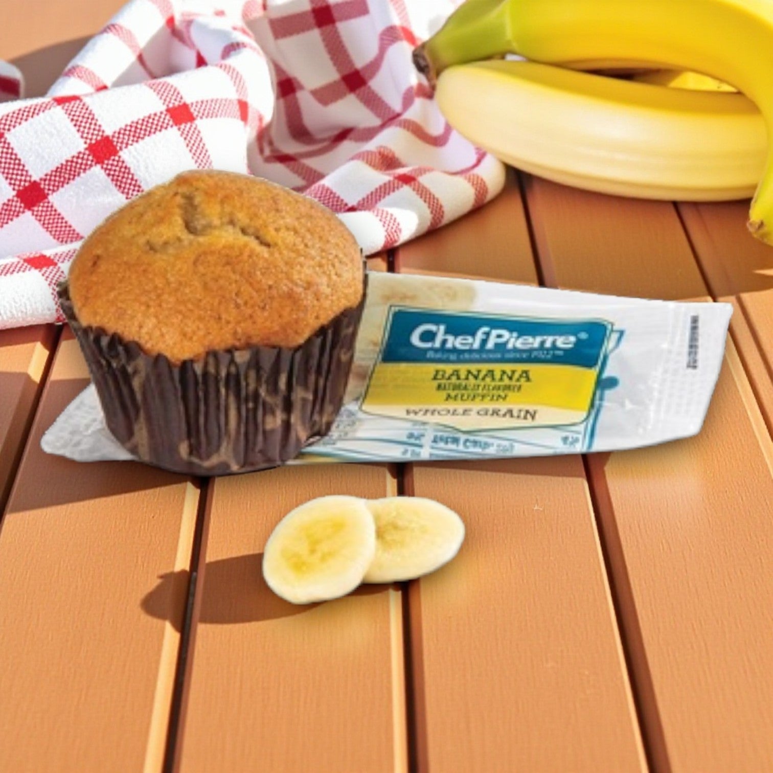 A Chef Pierre Whole Grain Banana Muffin, individually wrapped and 2 oz each, sits beside banana slices and a red-and-white cloth—perfect for an easy grab-and-go snack. Available in 48-count packs.