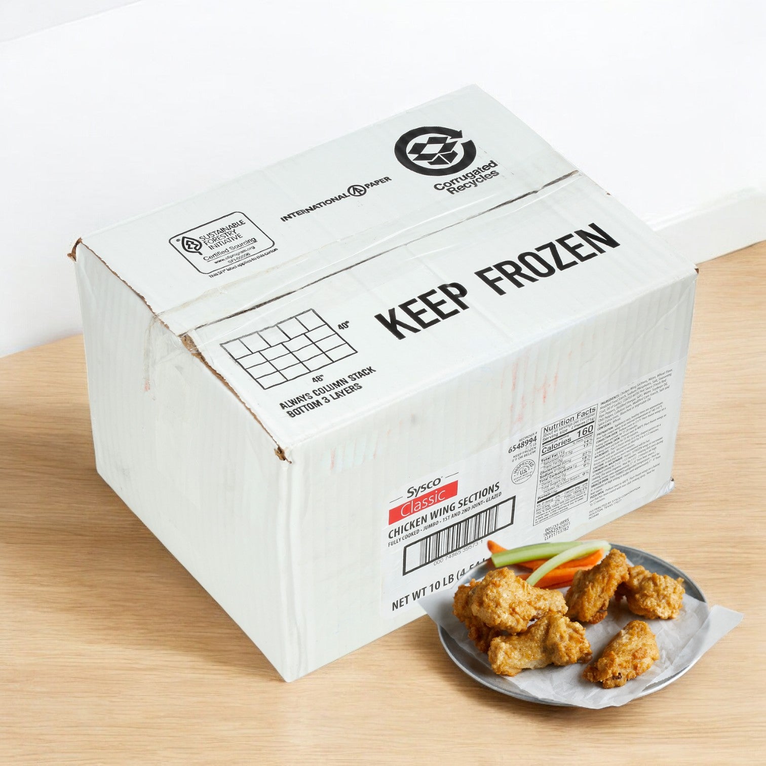 A "Keep Frozen" box containing Sysco Classic Chicken Wing Fire Pre Cooked 1st & 2nd Joints Large sits on a wooden surface near a plate of spicy wings, carrot sticks, and celery.