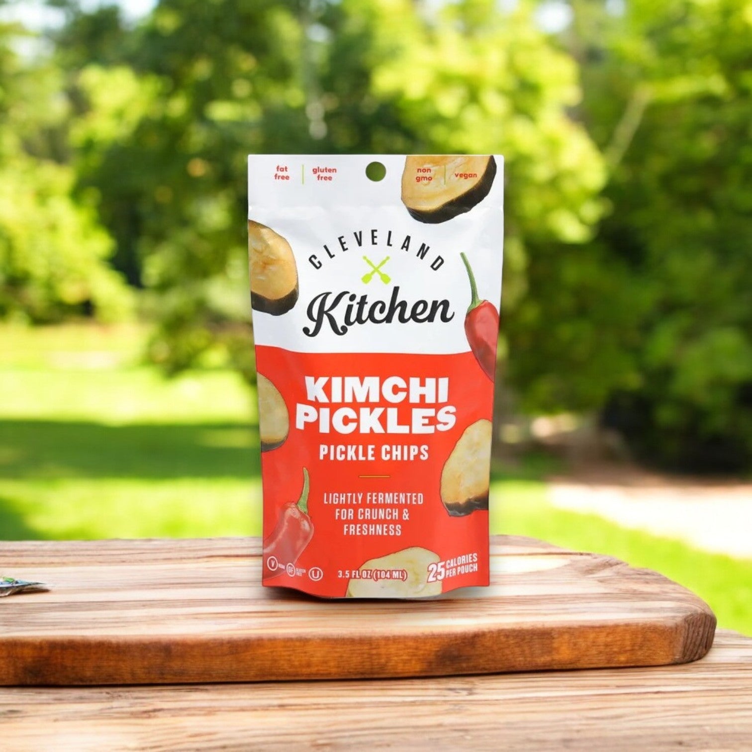 A 3.5 oz bag of Cleveland Kitchen Kimchi Pickle Chips sits on a wooden board outdoors, with greenery softly blurred in the background.