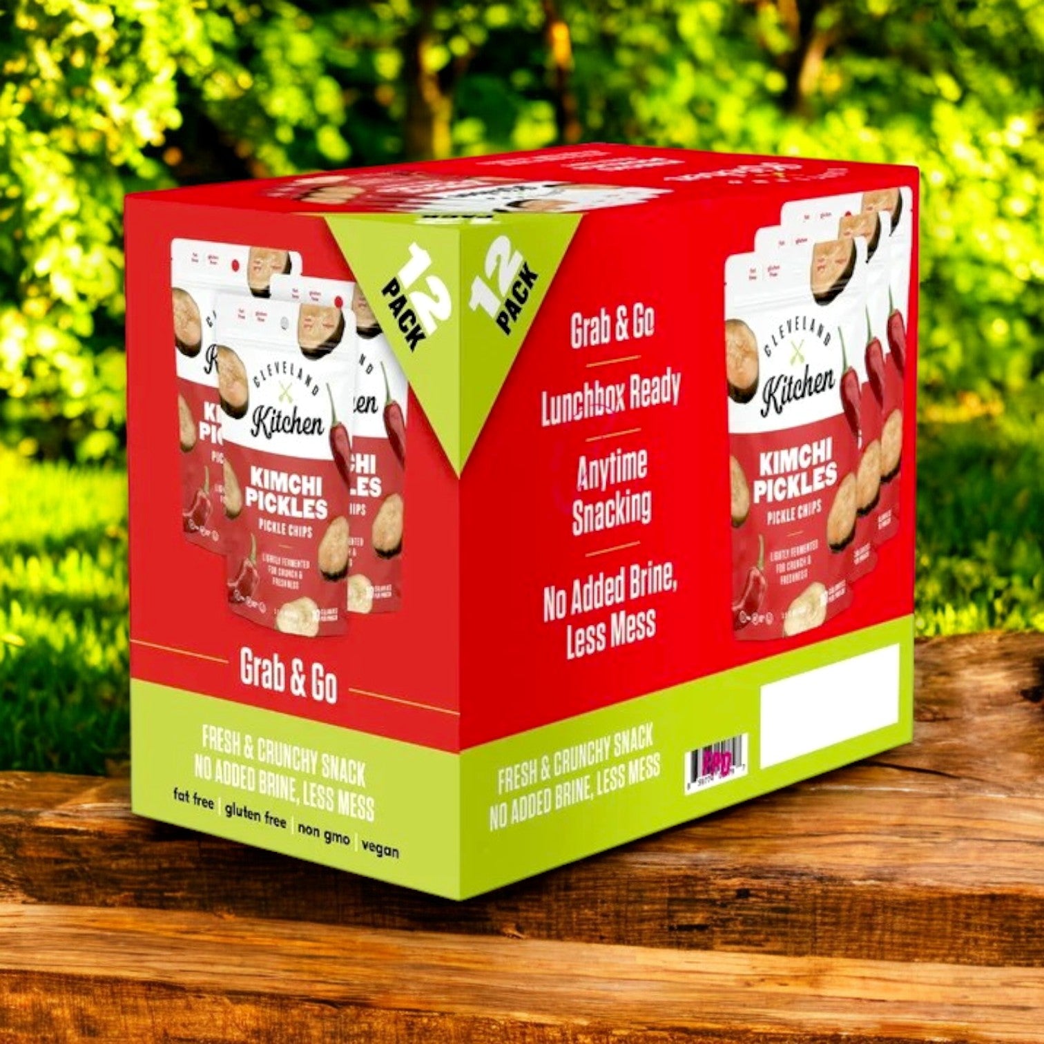 A 12-count box of Cleveland Kitchen Kimchi Pickle Chips (3.5 oz each) is displayed outdoors on wood with a lush green backdrop, showcasing these probiotic-rich snacks and their gut health benefits.