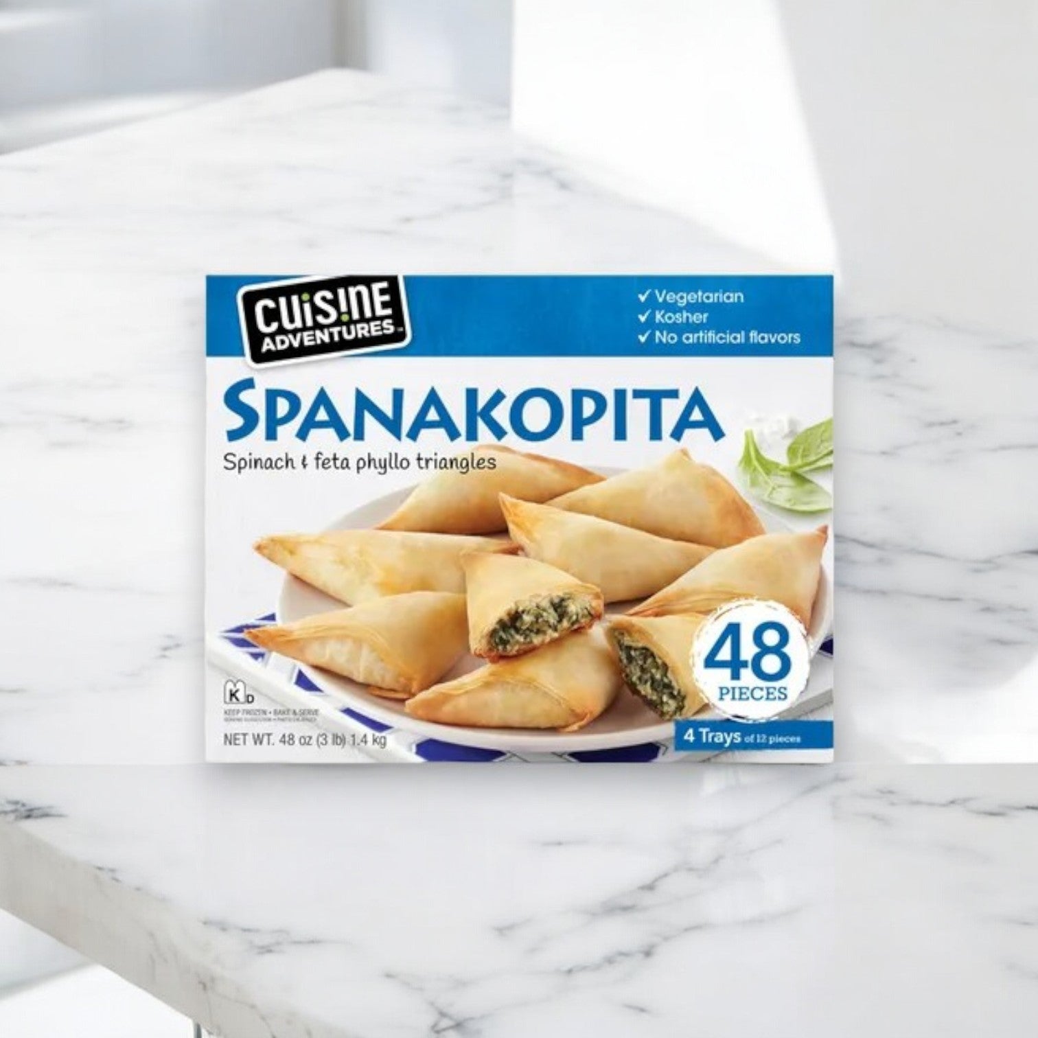 A set of Cuisine Adventure Spanakopita boxes, resting on marble, showcases spinach and feta phyllo triangles. The 48 oz package includes four boxes of vegetarian and kosher snacks made without artificial flavors, offering a Mediterranean culinary delight.
