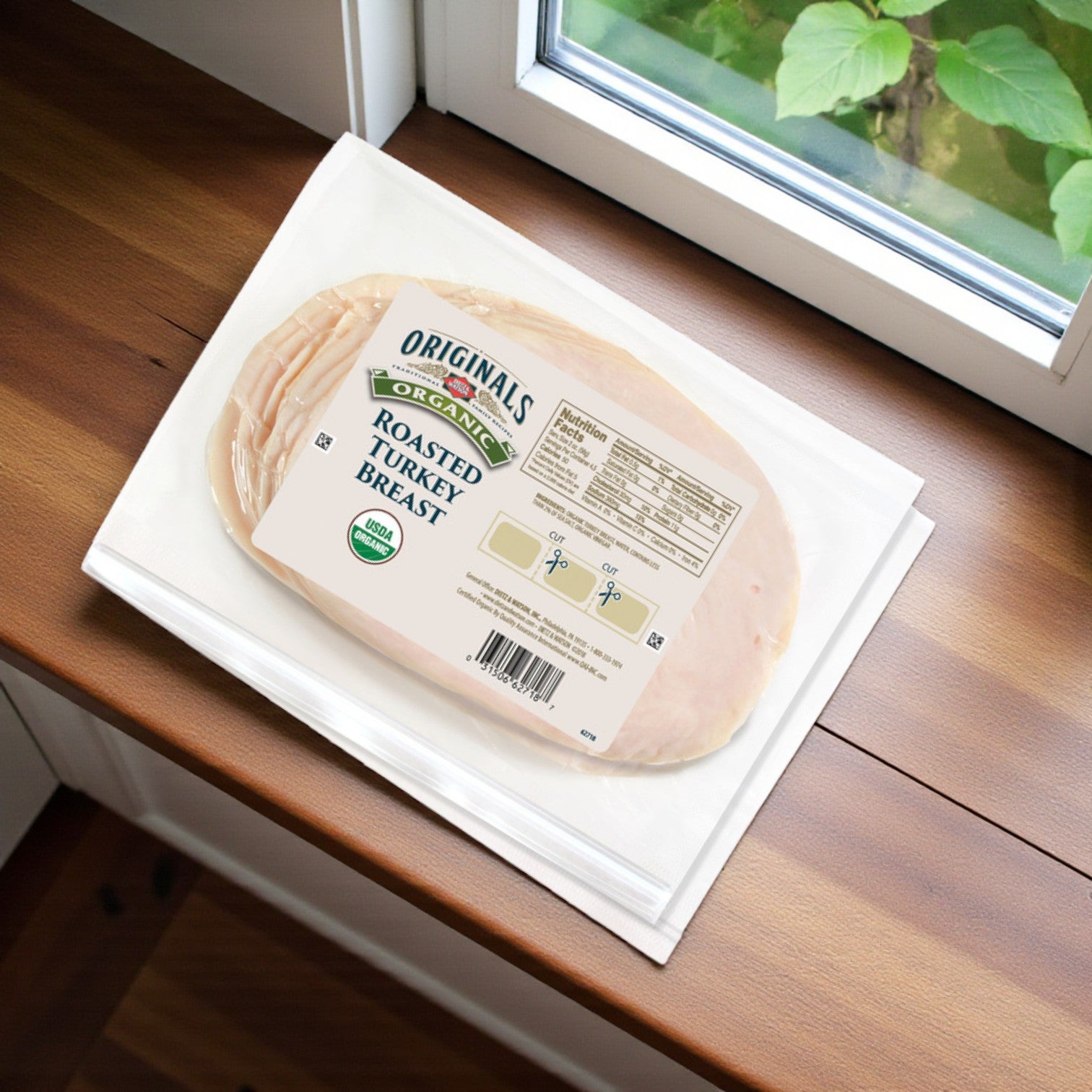 A package of Dietz & Watson Organic Roasted Turkey Breast – 9 oz, 3 Count rests on a stack of white napkins atop a wooden countertop near a window.