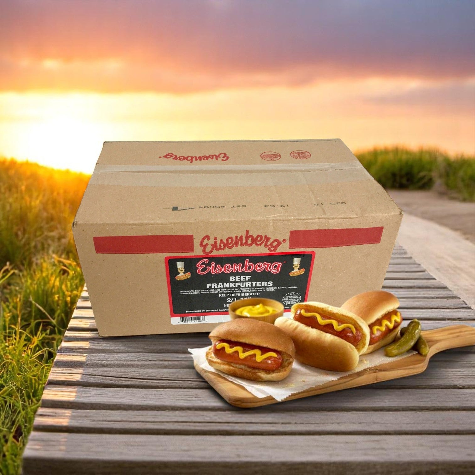A box labeled "Easy Lunches Eisenberg Mini Angus Beef Franks – 16/1 Size (4 Packs of 3 lbs Each)" sits on a wooden surface outdoors, with three mini beef franks and pickles on a cutting board, sunset and grass in the background.