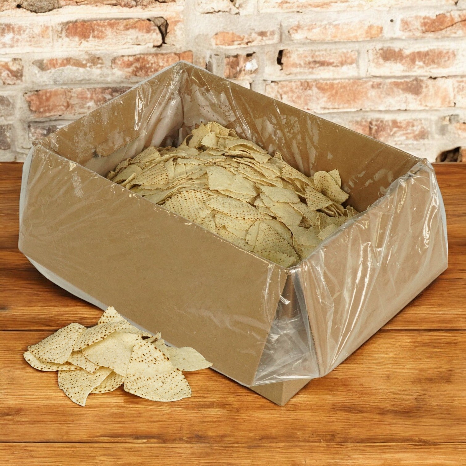 A 20-pound bulk pack of El Milagro White Corn Tortillas chips, irregular in shape and light-colored, spills from an open cardboard box onto a wooden surface against a rustic brick wall. The chips have a classic 4-way cut reminiscent of white corn tortillas.