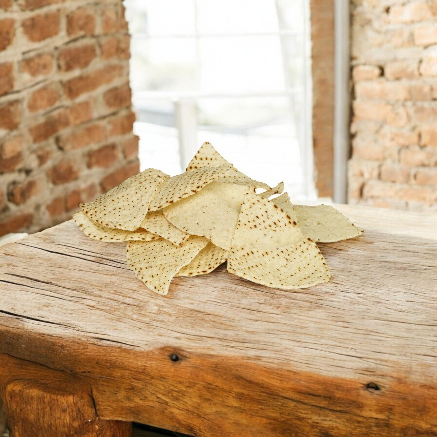 A rustic wooden table features white corn tortillas from El Milagro, presented as a pile of tortilla chips. The background's brick walls and window highlight that these chips come from the expansive El Milagro 20-pound bulk pack.