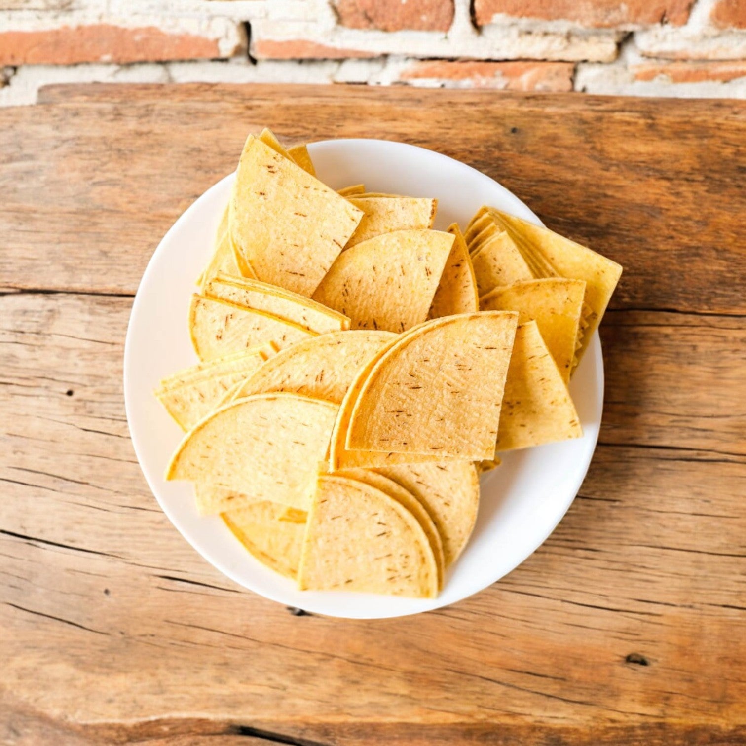 Triangular chips from El Milagro White Corn Tortillas are arranged on a wooden surface, set against a rustic brick wall.