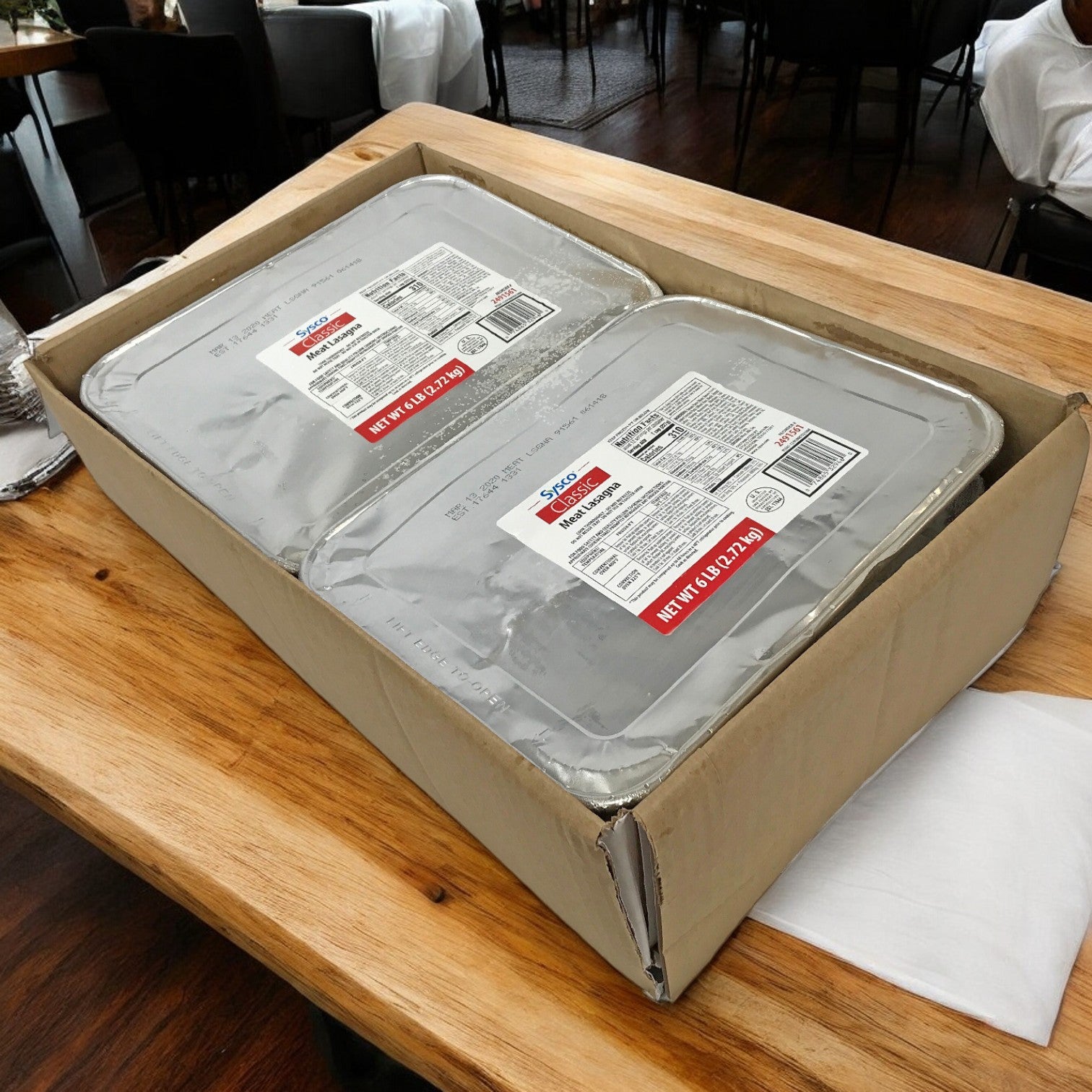 On a wooden table sits a cardboard box containing two large sealed Sysco Classic metal trays labeled as meat lasagna, promising layers of cheese and savory meat sauce.