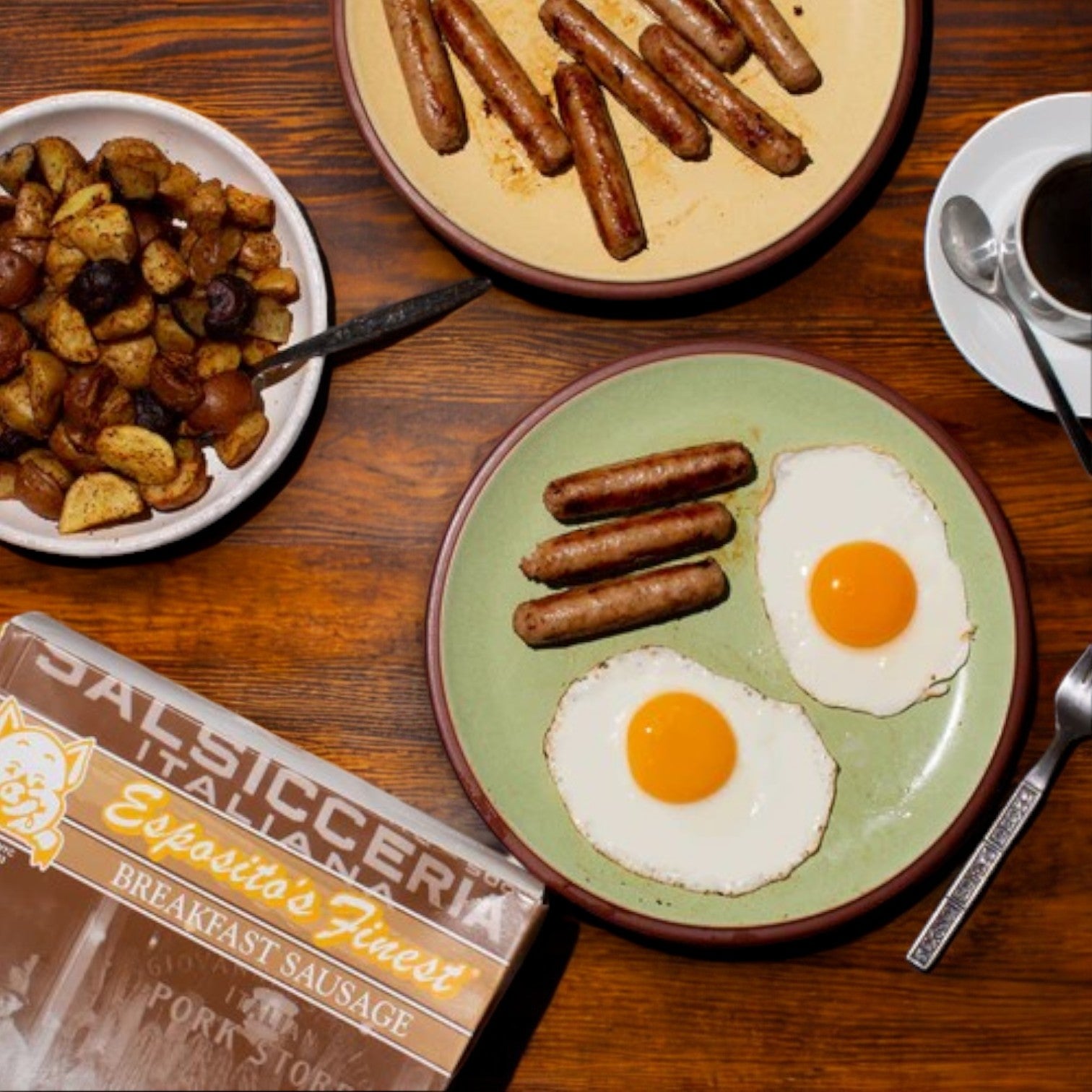 Plates holding cooked breakfast sausage links, two fried eggs, and roasted potatoes rest on a wooden table. A box labeled "Esposito's Finest Quality Sausage – Breakfast Sausage" promises an authentic Italian recipe with premium quality pork. Nearby, a steaming cup of coffee completes the scene.