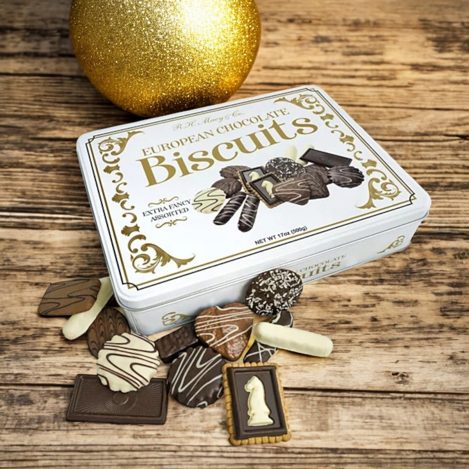A beautifully displayed R.H. Macy & Co. European Chocolate Biscuit Tin by Macy's is open, revealing an assortment of exquisitely shaped and decorated treats scattered across a wooden surface. In the background, a large gold ornament enhances the scene with an added touch of luxury.