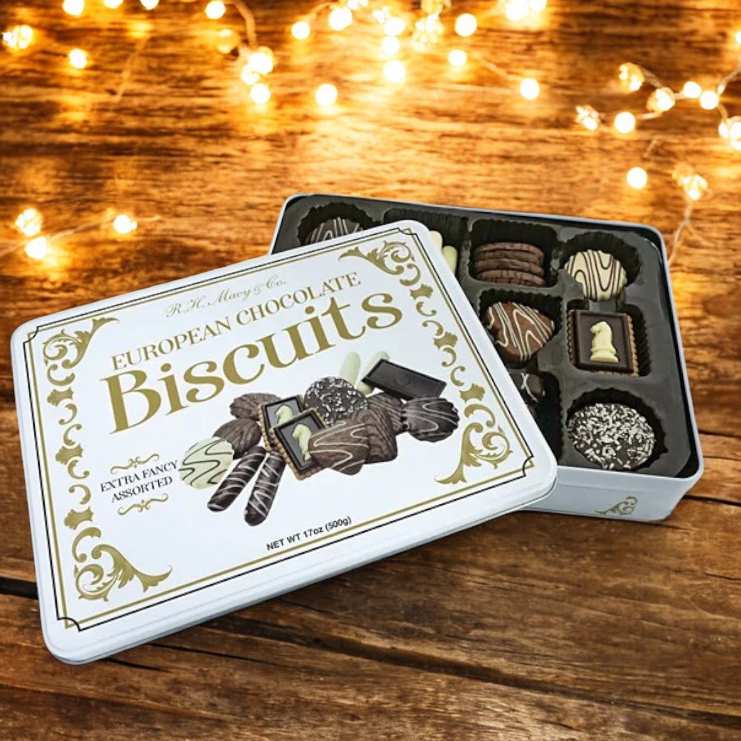 An open tin of R.H. Macy & Co. European Chocolate Biscuit Tin from Macy's, filled with a variety of premium chocolate cookies, is showcased. The elegant packaging is beautifully displayed against a wooden surface adorned with string lights.