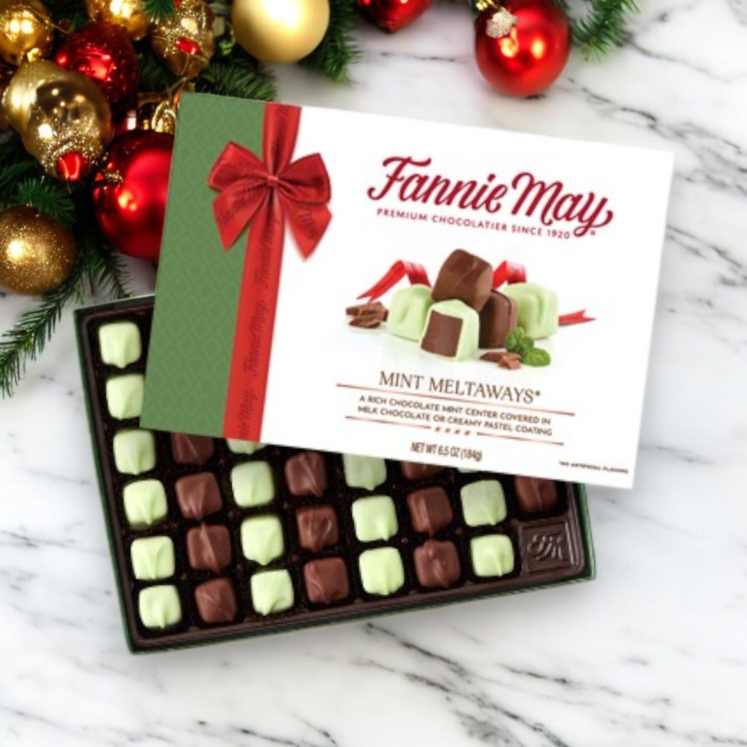 An open box of Fannie May Candy, Mint Meltaways, 1 Count showcases a tempting selection of green and brown mint chocolate squares, beautifully complemented by festive ornaments.
