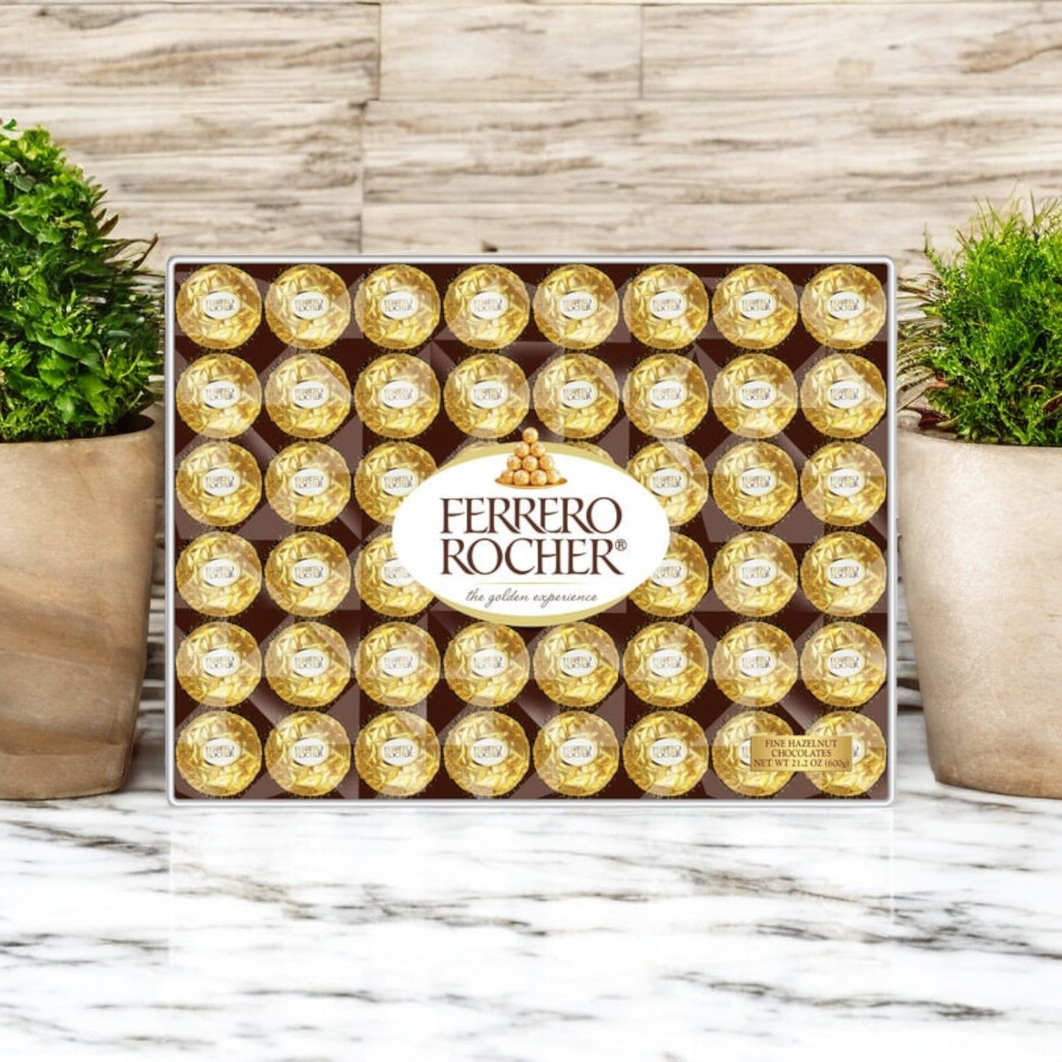 A Ferrero Rocher Milk Chocolate Hazelnut Candy 21.2 oz, 48 Count gift box by Ferrero Rocher is displayed on a marble surface between two potted green plants.