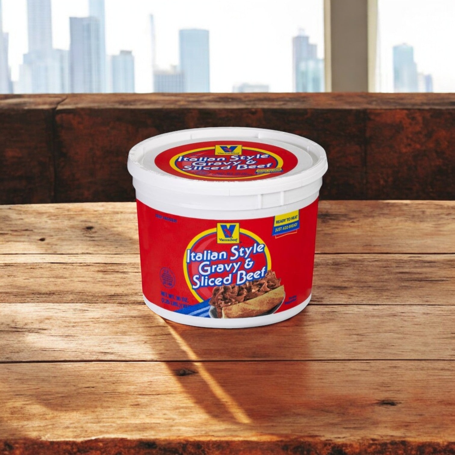 A tub of Easy Lunches Chicago’s Best Combo Kit – Lou Malnati’s Deep Dish & Vienna Italian Beef sits on a wooden table, ready to elevate your meal, with a city skyline visible through large windows in the background.