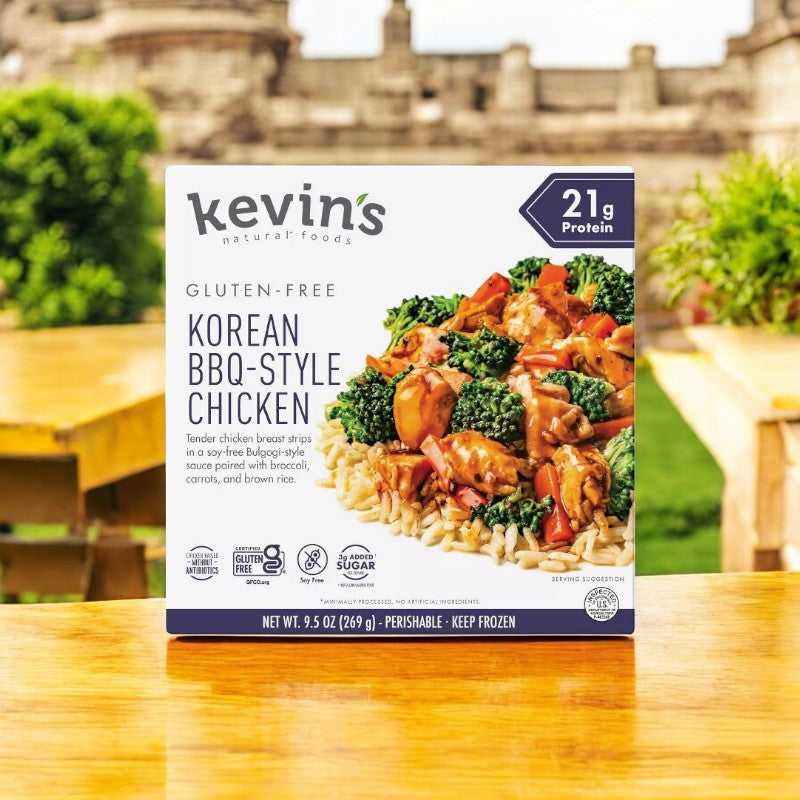 A box of Kevin's Natural Foods Korean BBQ-Style Chicken - 1 Count by Kevin's rests on a wooden table outdoors, with greenery and stone structures blurred in the background.