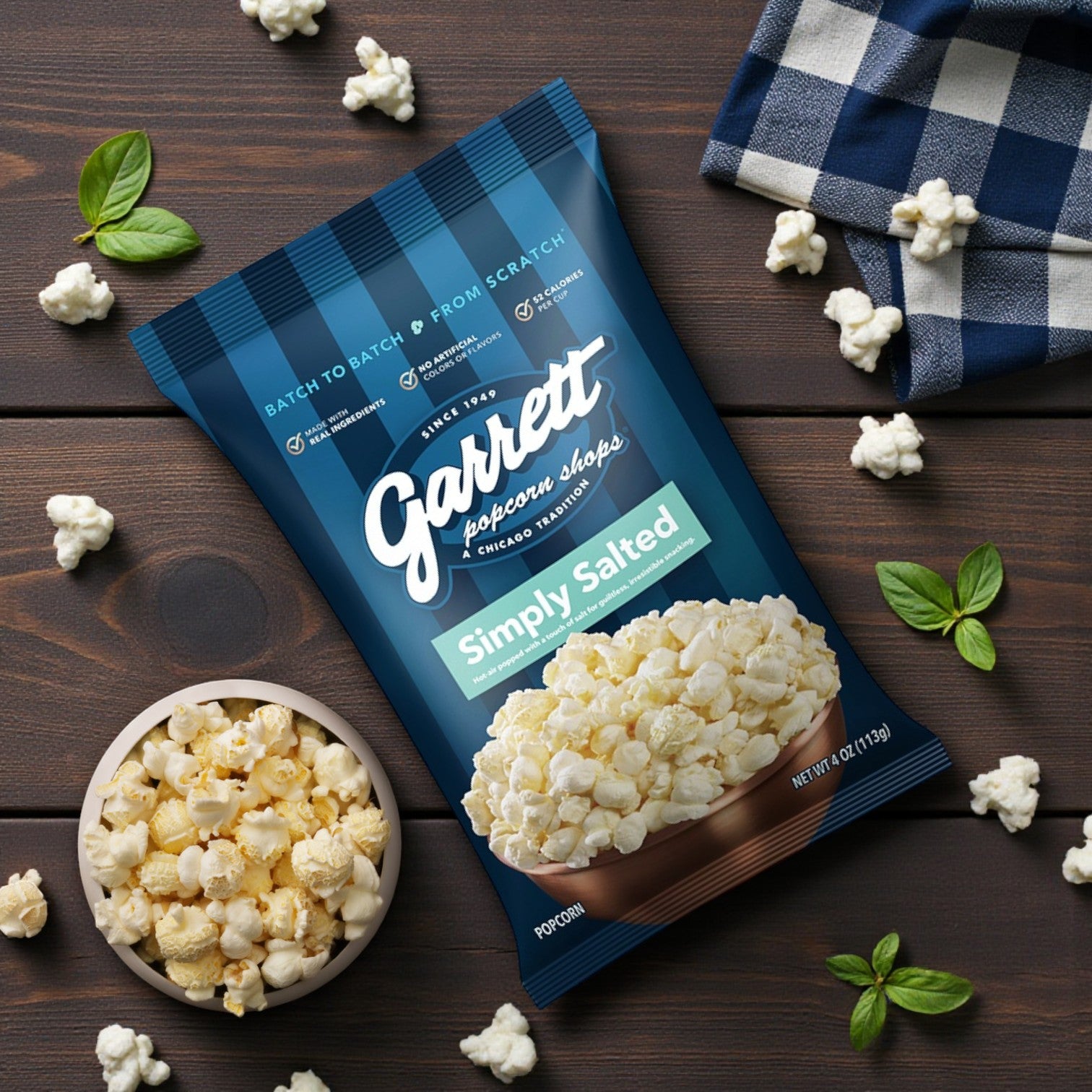 A 4 oz bag of Garrett Popcorn – Simply Salted by Garrett rests on a wooden surface with a bowl of popcorn, scattered kernels, basil leaves, and a blue checkered cloth.