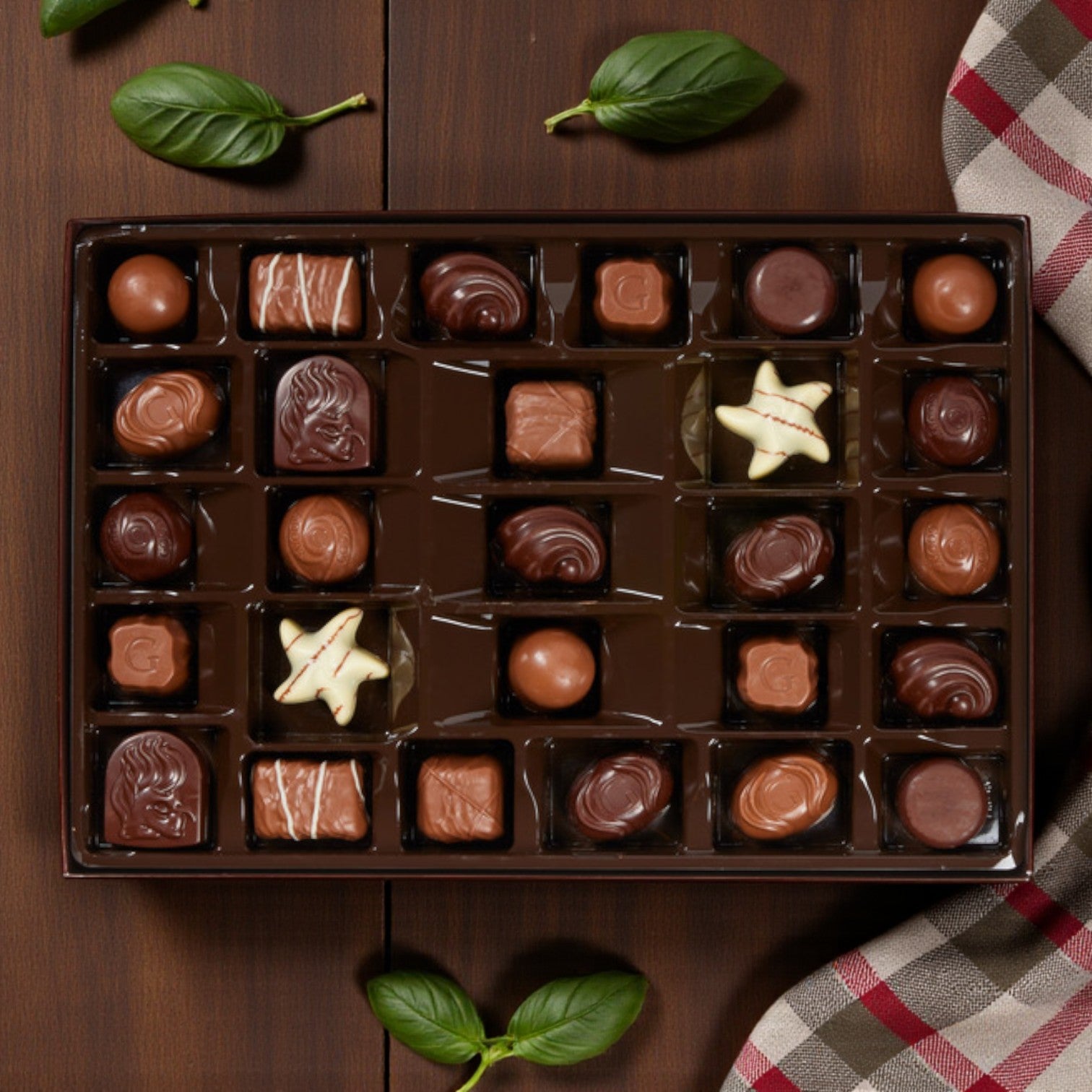 A Godiva Premium Assorted Chocolates – 27 Count gift box is open on a wooden surface, with green basil leaves and a plaid cloth nearby.