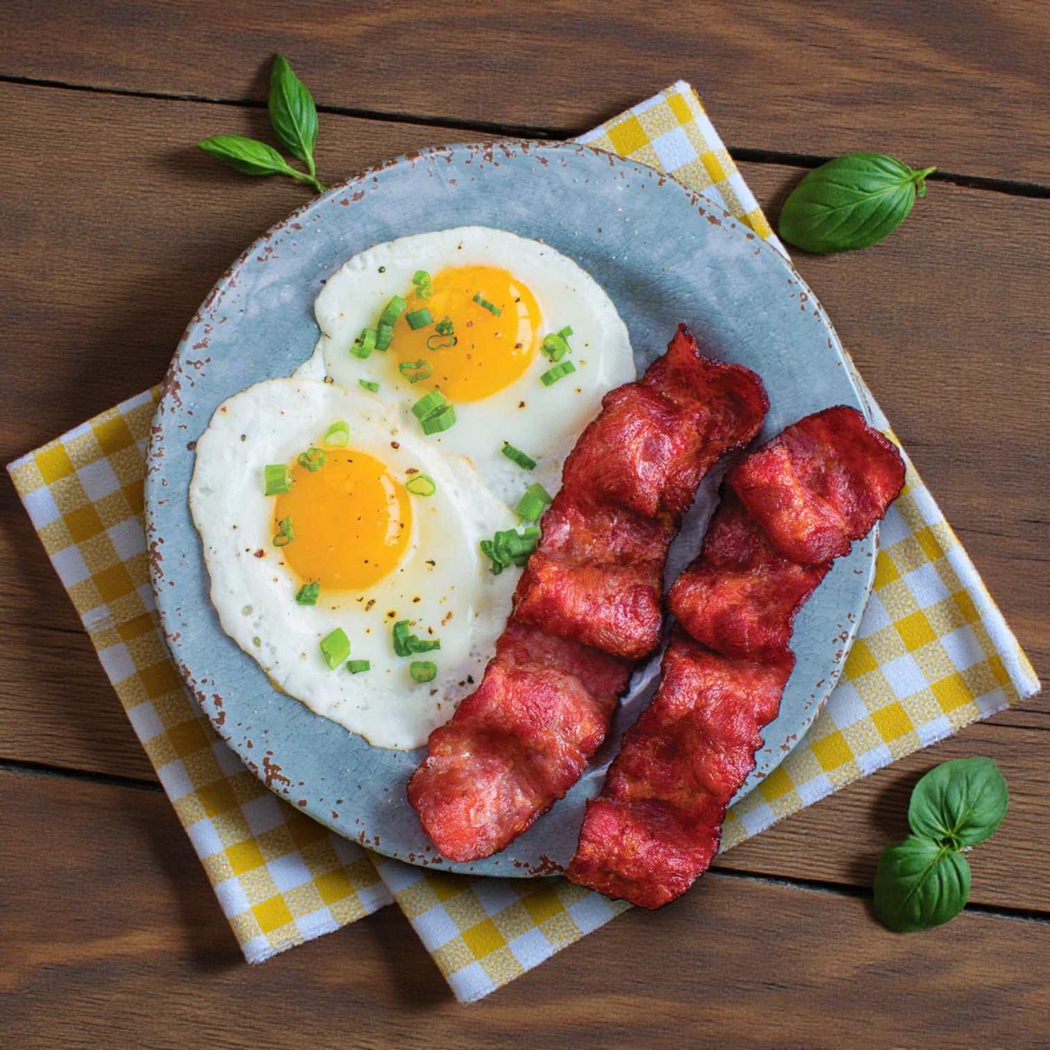 A plate with two sunny-side up eggs topped with green onions and three strips of Godshall's Uncured Fully-Cooked Turkey Bacon (from the 40 oz 3 Pack Bundle), served on a checkered napkin on a wooden table.