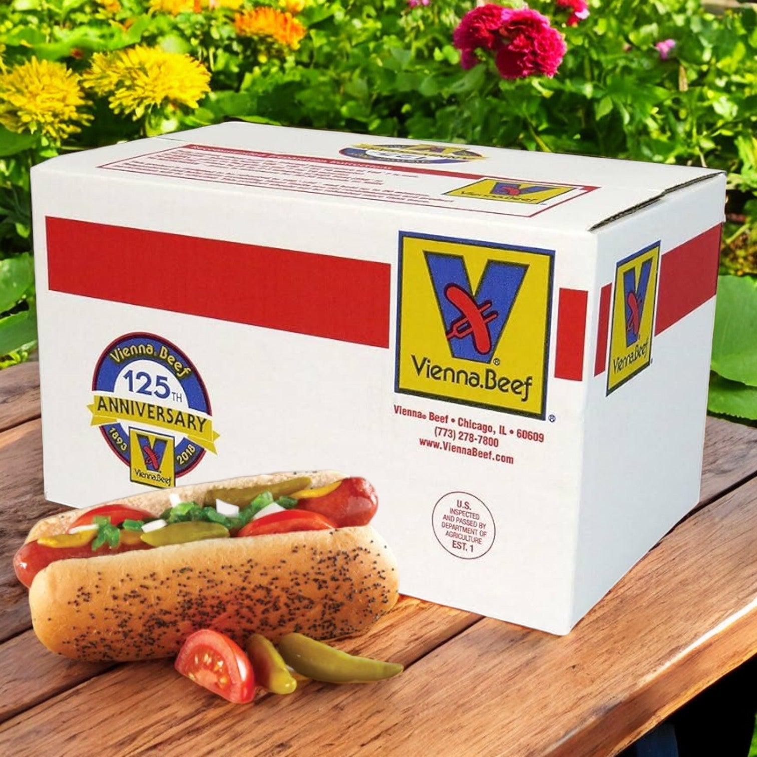 A Vienna Beef All-Beef 5:1 Hot Dogs box (2 bags, 5 lbs each—50 hot dogs) sits on a wooden table outdoors, with a classic Chicago-style all-beef hot dog, sliced tomato, and pickled peppers in front; bright flowers in the background.