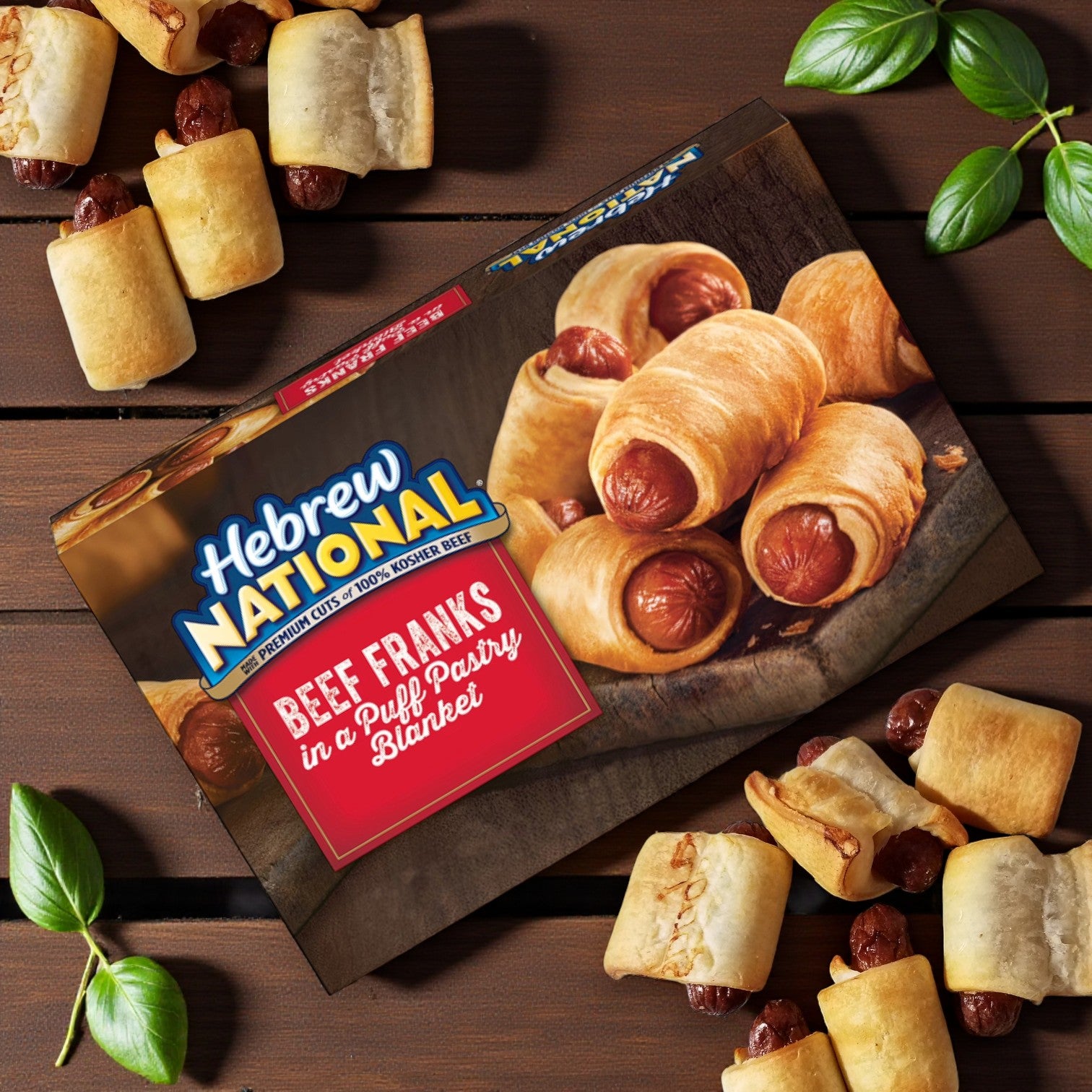 An 18 oz box of Hebrew National Beef Franks in a Puff Pastry Blanket rests on a wooden table with golden pastry pieces and fresh basil leaves—bite-sized kosher beef franks ideal for any gathering.