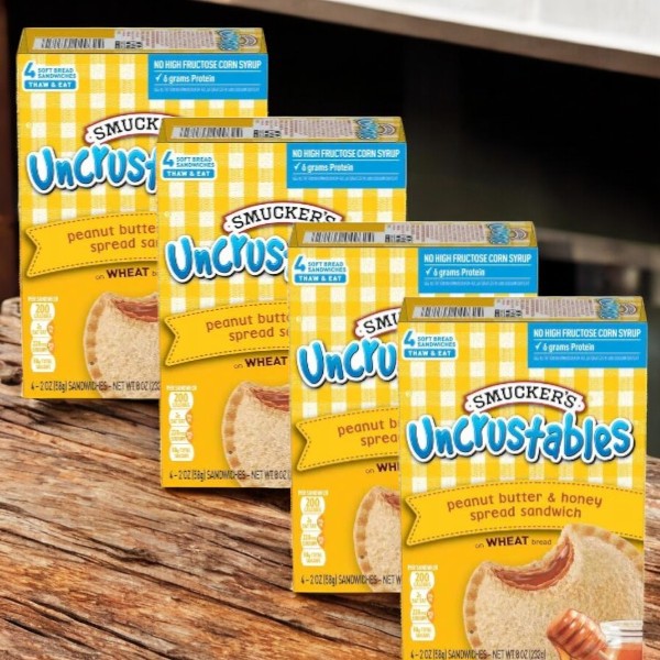 Four boxes of Uncrustables Peanut Butter & Honey Spread Sandwiches (16 sandwiches total by Uncrustable Smuckers) are displayed on a wooden surface.