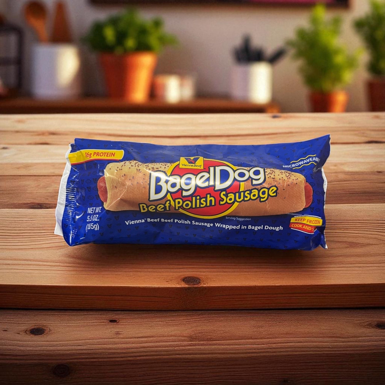 A packaged Vienna Beef Polish Sausage Bagel Dog sits on a wooden table with a kitchen background. The packaging features Vienna Beef branding and highlights high protein, freezer-to-oven prep, and premium hot dog bun taste.