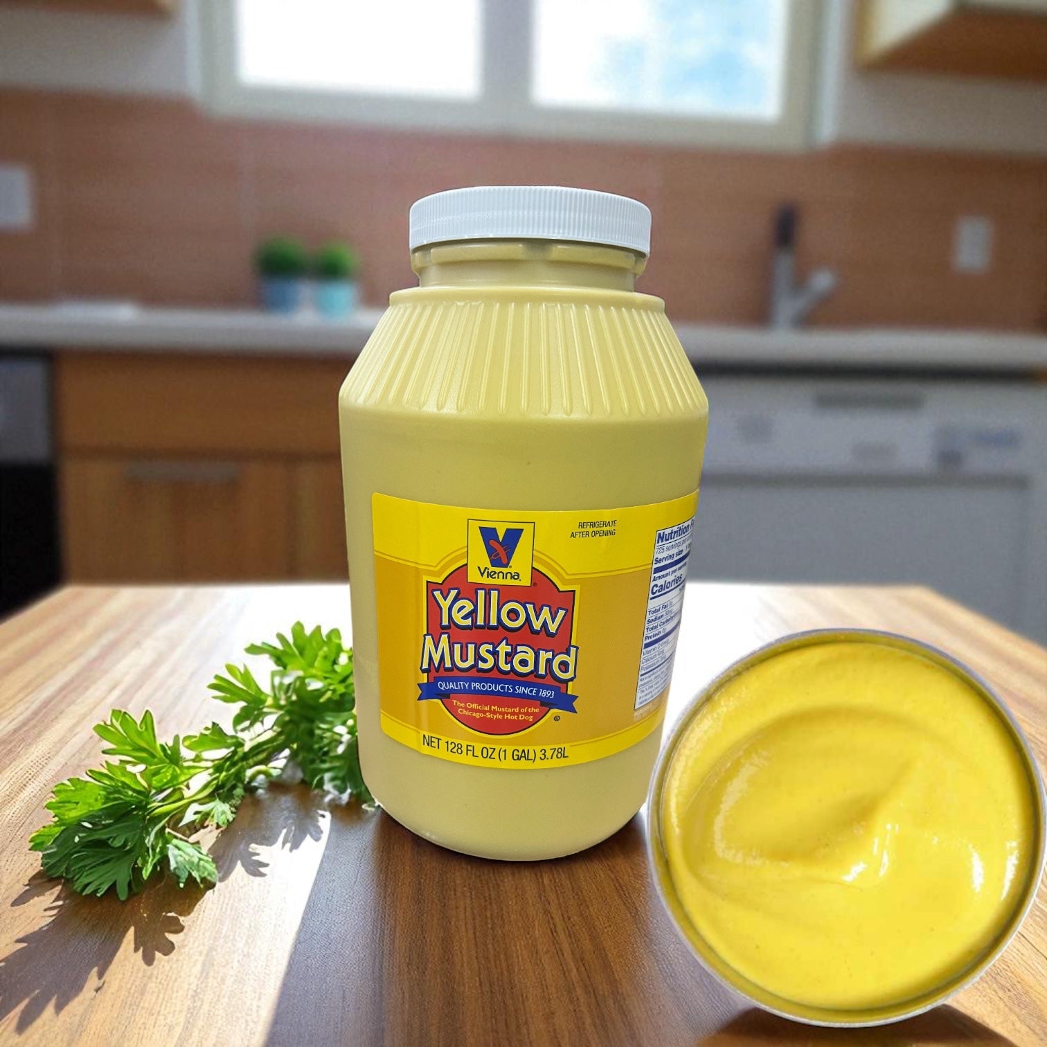 A 1-gallon jar of Vienna Beef Vienna Yellow Mustard sits on a wooden kitchen table next to a small bowl of mustard and a sprig of parsley, with a kitchen in the background.