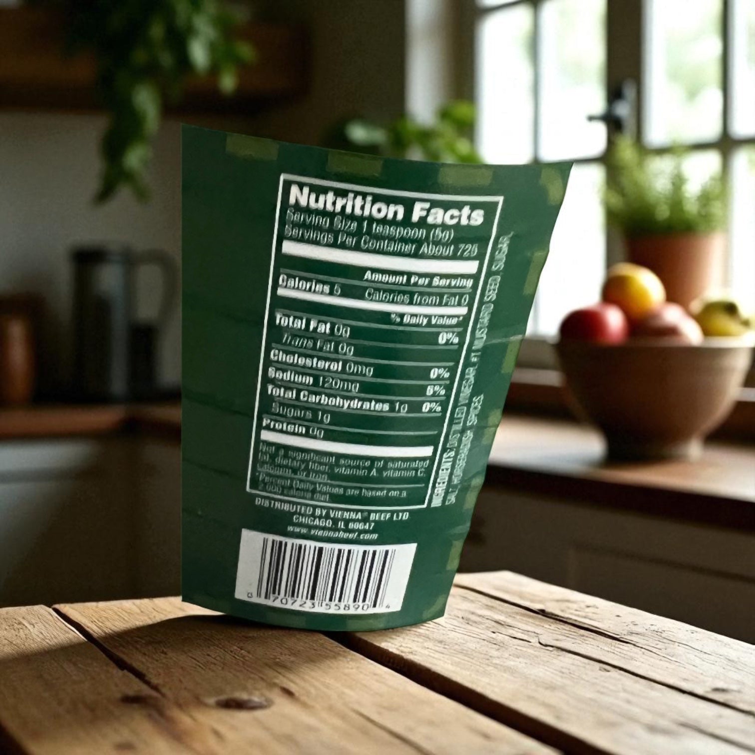 A jar of Vienna Beef Vienna Horse Radish Mustard 1 Gallon sits on a kitchen counter, next to a green food package with nutrition facts, with fruit and a window blurred in the background.