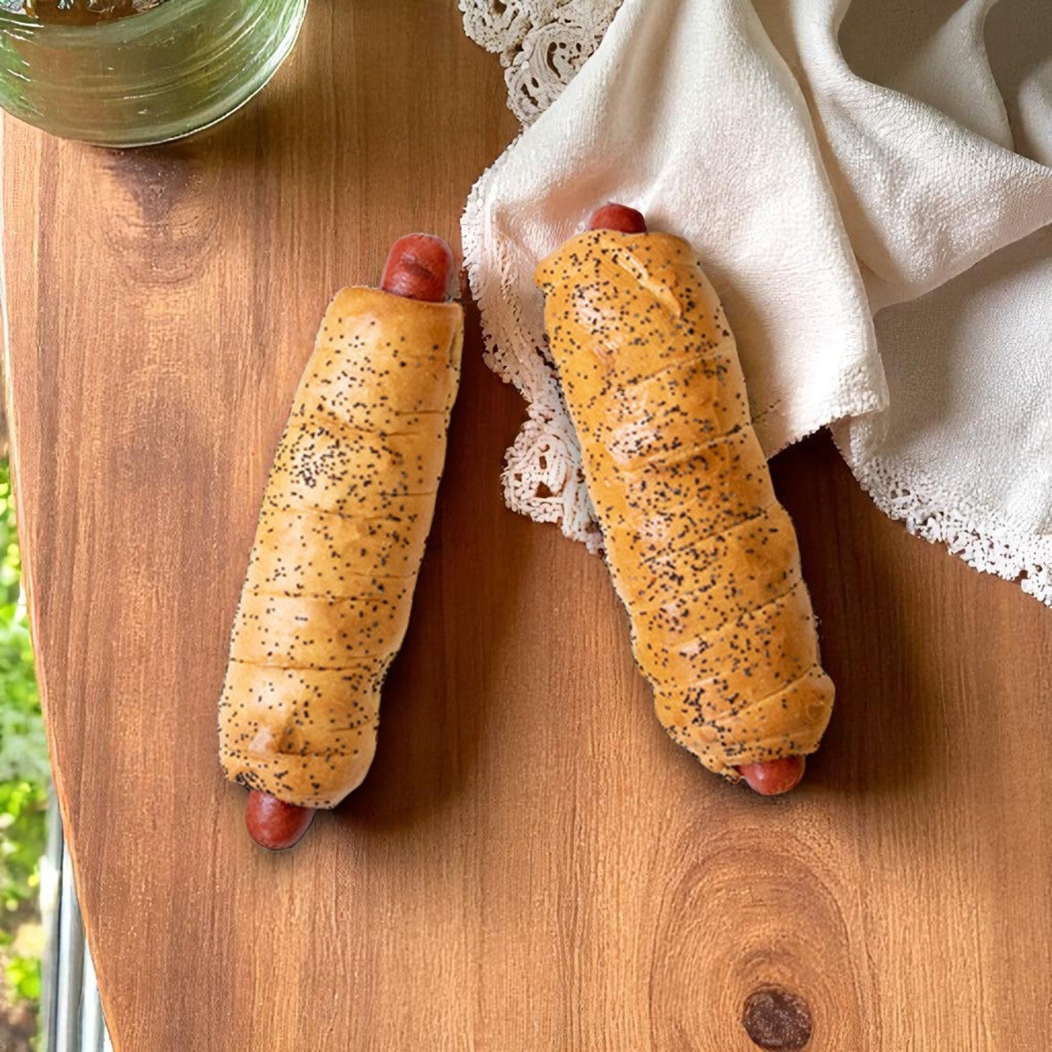 Two Vienna Beef BagelDog – 6.5 oz. hot dogs sit on a wooden surface by a white lace cloth and a glass of water, offering the classic Chicago flavor in every bagel-wrapped bite. Available in packs of 24 from Vienna Beef.