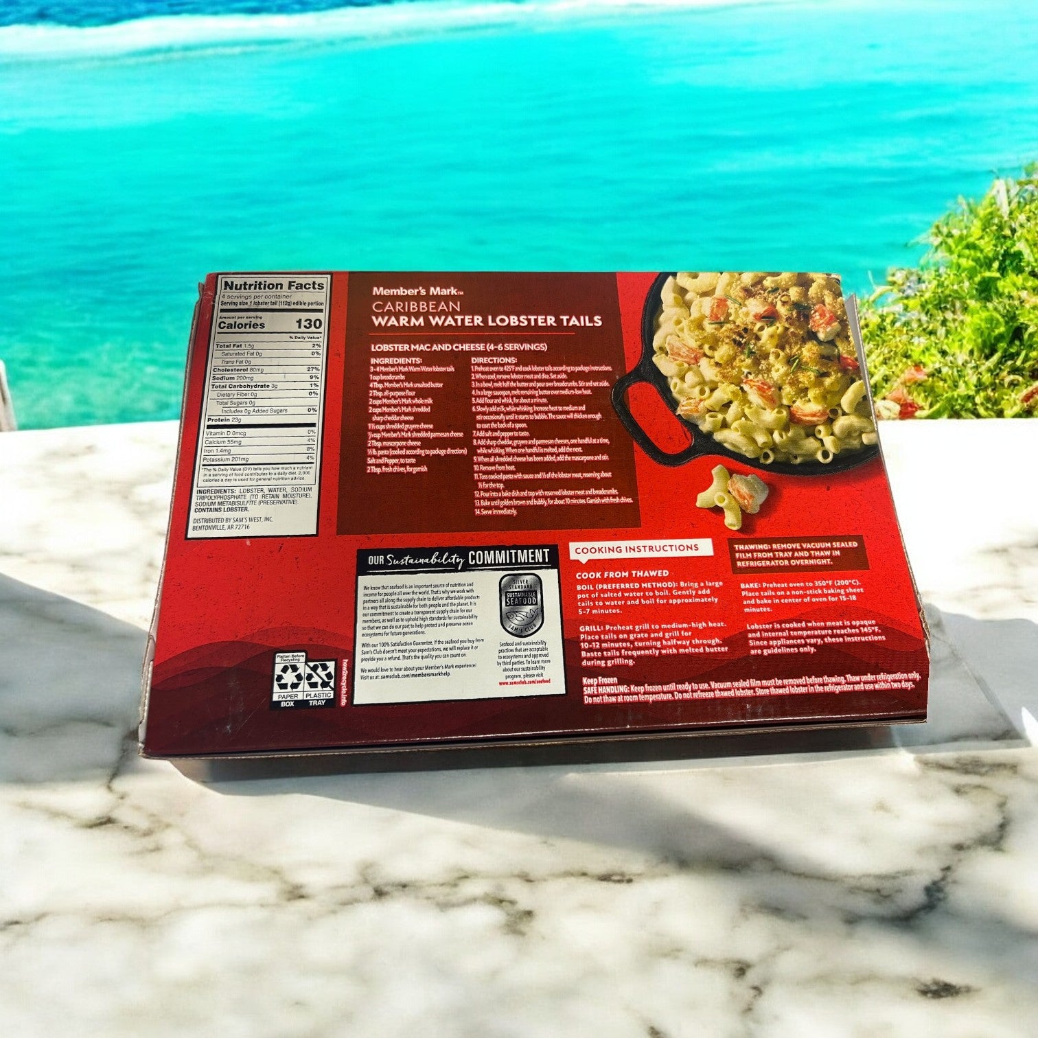 A box of Easy Lunches Member's Mark Warm Water Lobster Tails, Frozen, 4 Count-3 Pack rests on a marble surface with a blue ocean and greenery in the background.