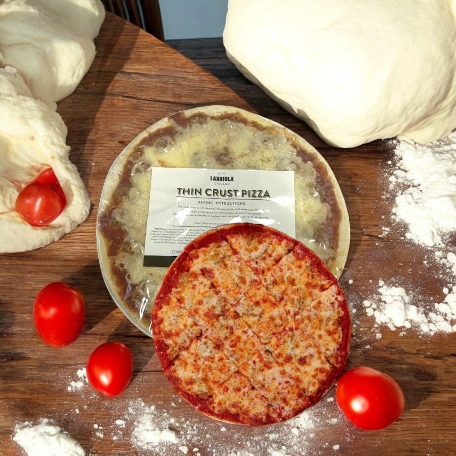 A Labriola Chicago Sausage Thin Crust Pizza-12inch-1 Count sits on a wooden table with raw dough and flour-dusted tomatoes, adorned with savory mozzarella cheese.
