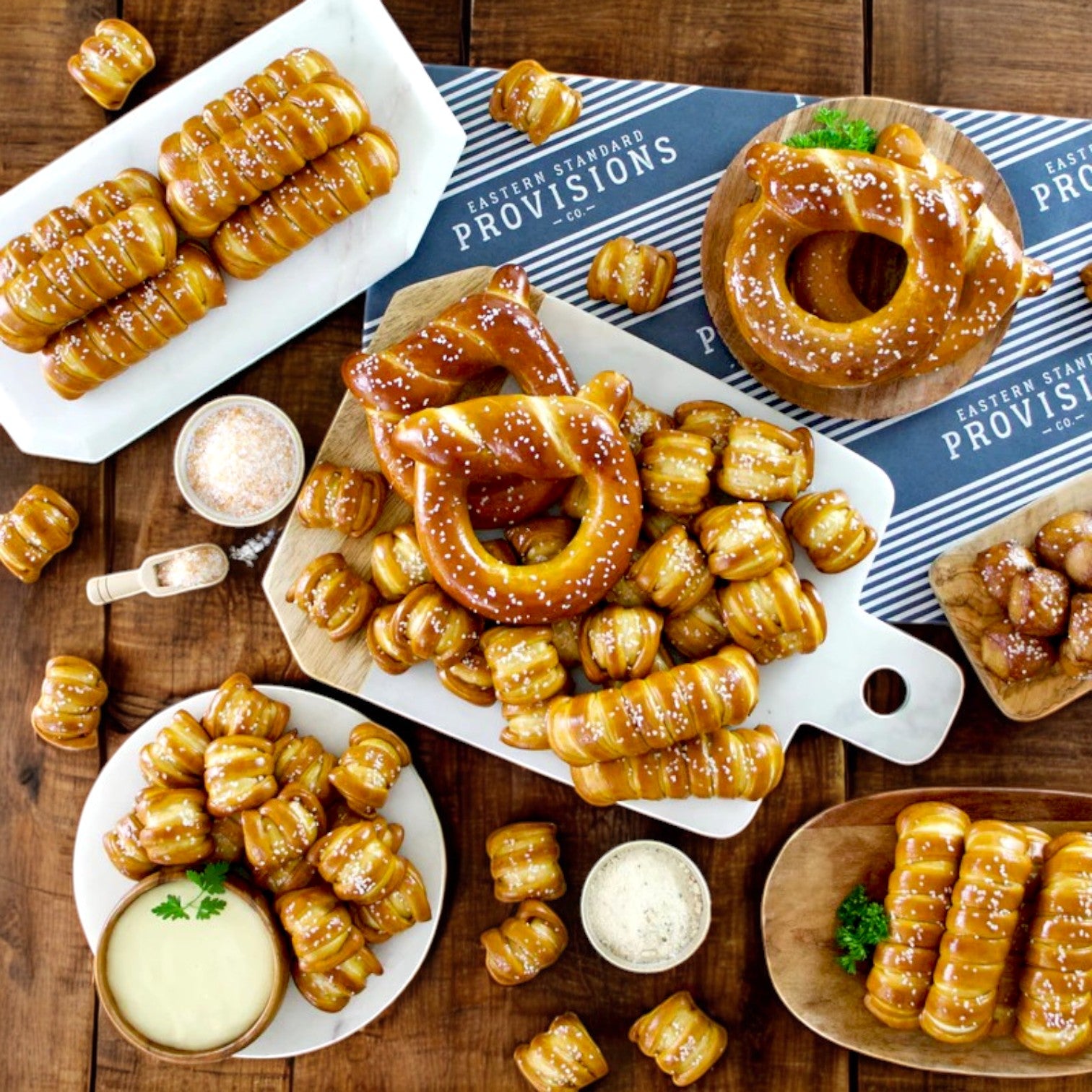 Assorted artisanal soft pretzels in various shapes and salted toppings are on a wooden table with dipping sauces. A cloth displays "Eastern Standard Provisions Pretzel 3-Pack Combo," highlighting favorites like the three cheese classic.