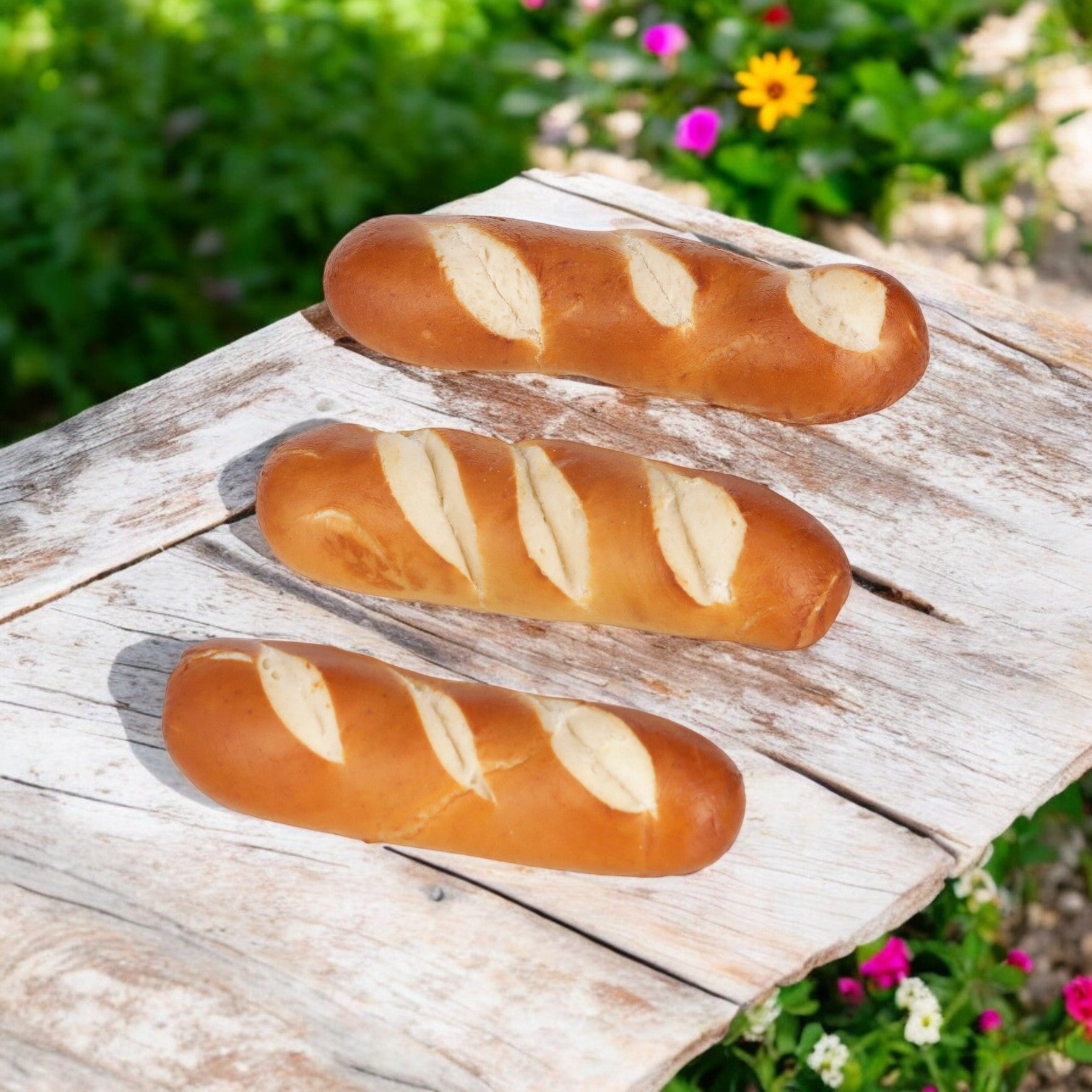 Three freshly baked baguettes with diagonal slashes lay on a rustic wooden table surrounded by grass and colorful flowers. Nearby, a basket of J&j Snacks' Pretzel Bavarian Stick Minis, 1.2 oz.-100 Count, entices with its golden hue, perfect for satisfying snack cravings.
