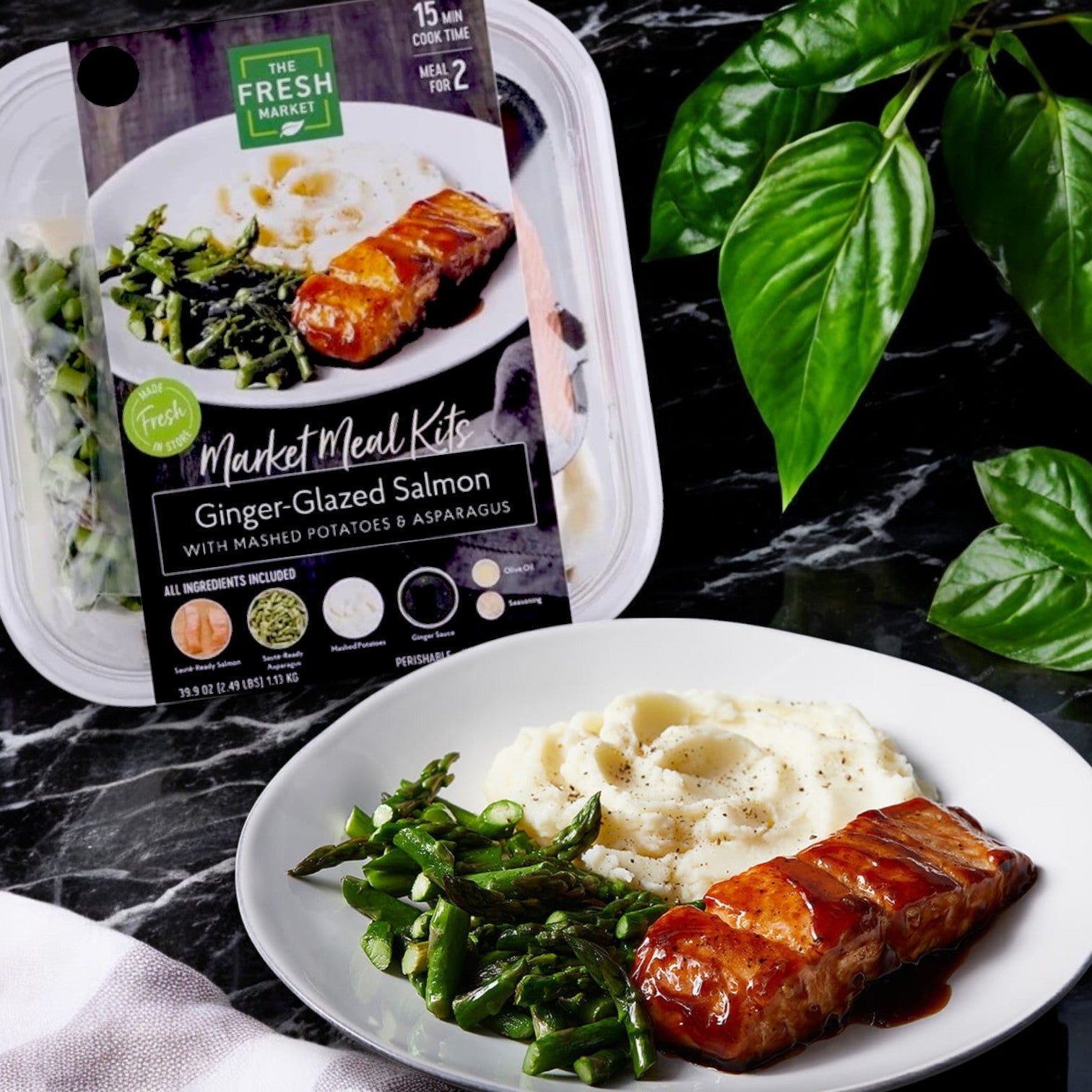 The Fresh Market's 40 oz Ginger Glazed Salmon Market Meal Kit sits beside a prepared plate of succulent salmon, creamy mashed potatoes, and fresh asparagus, with a leafy green plant enhancing the scene.