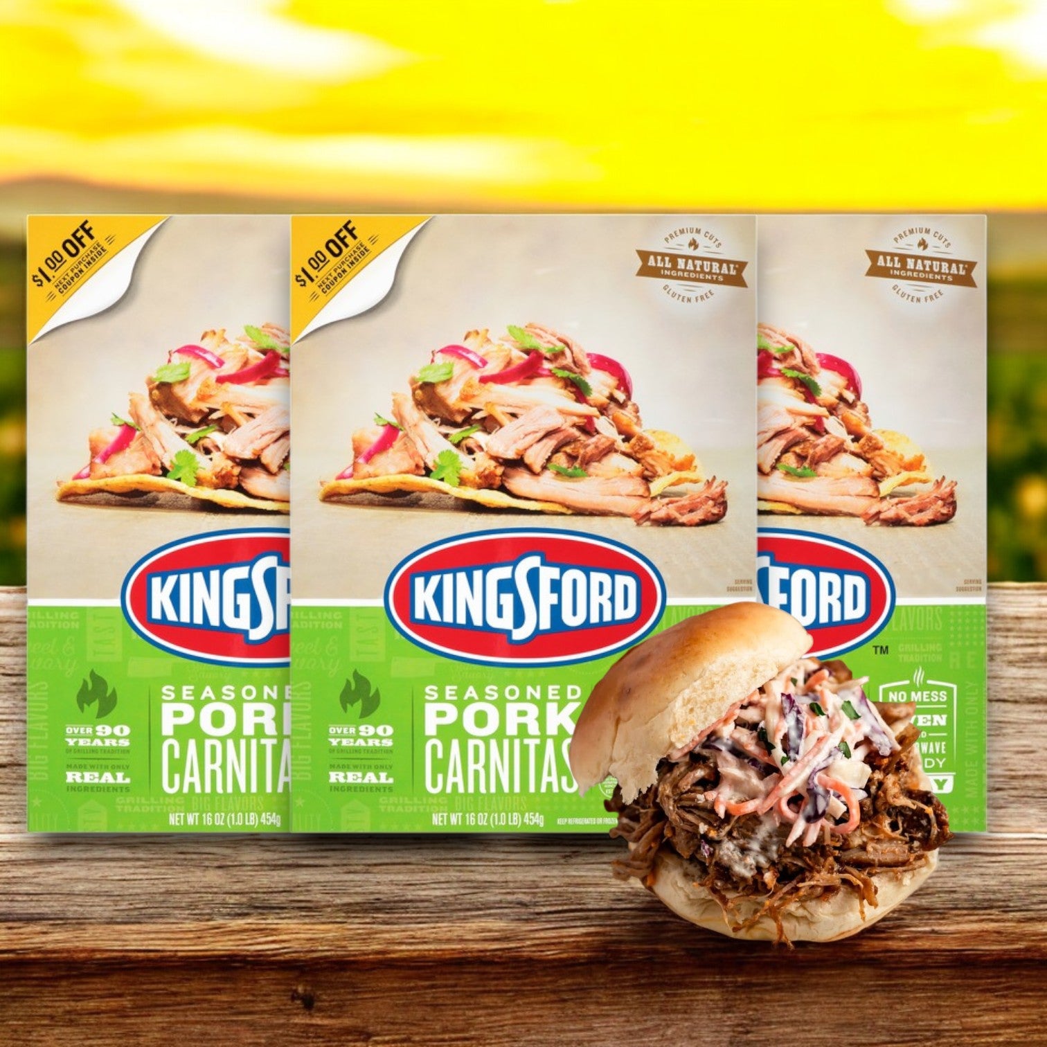 In front of a sunny landscape sits a box of Kingsford Seasoned Pork Carnitas – 16 oz -3 Count, perfectly slow-cooked and ready, with a sandwich showcasing the smoky-seasoned pork.