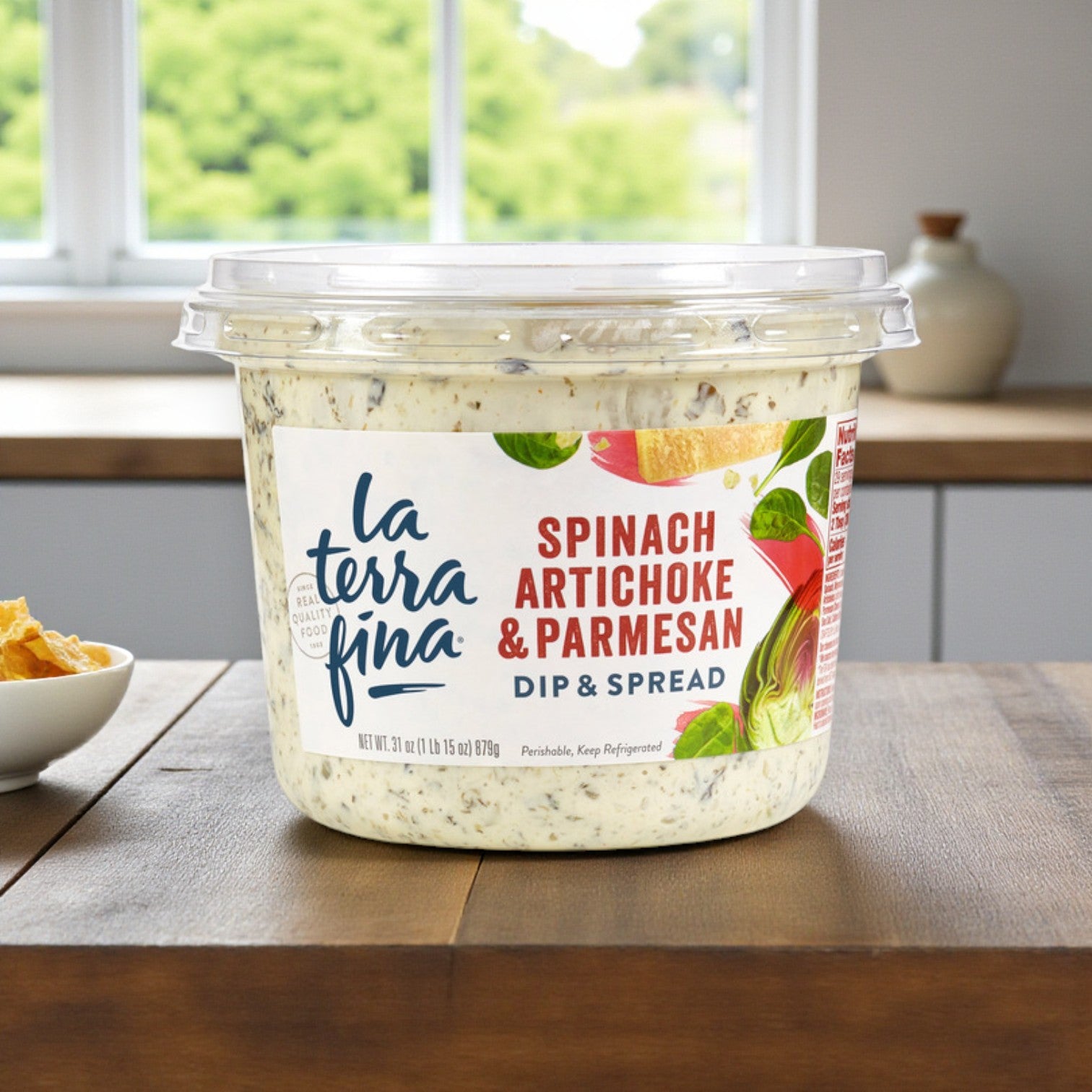 A 31 oz container of La Terra Fina Spinach, Artichoke & Parmesan Dip sits on the kitchen counter beside a bowl of chips, ready to enjoy.