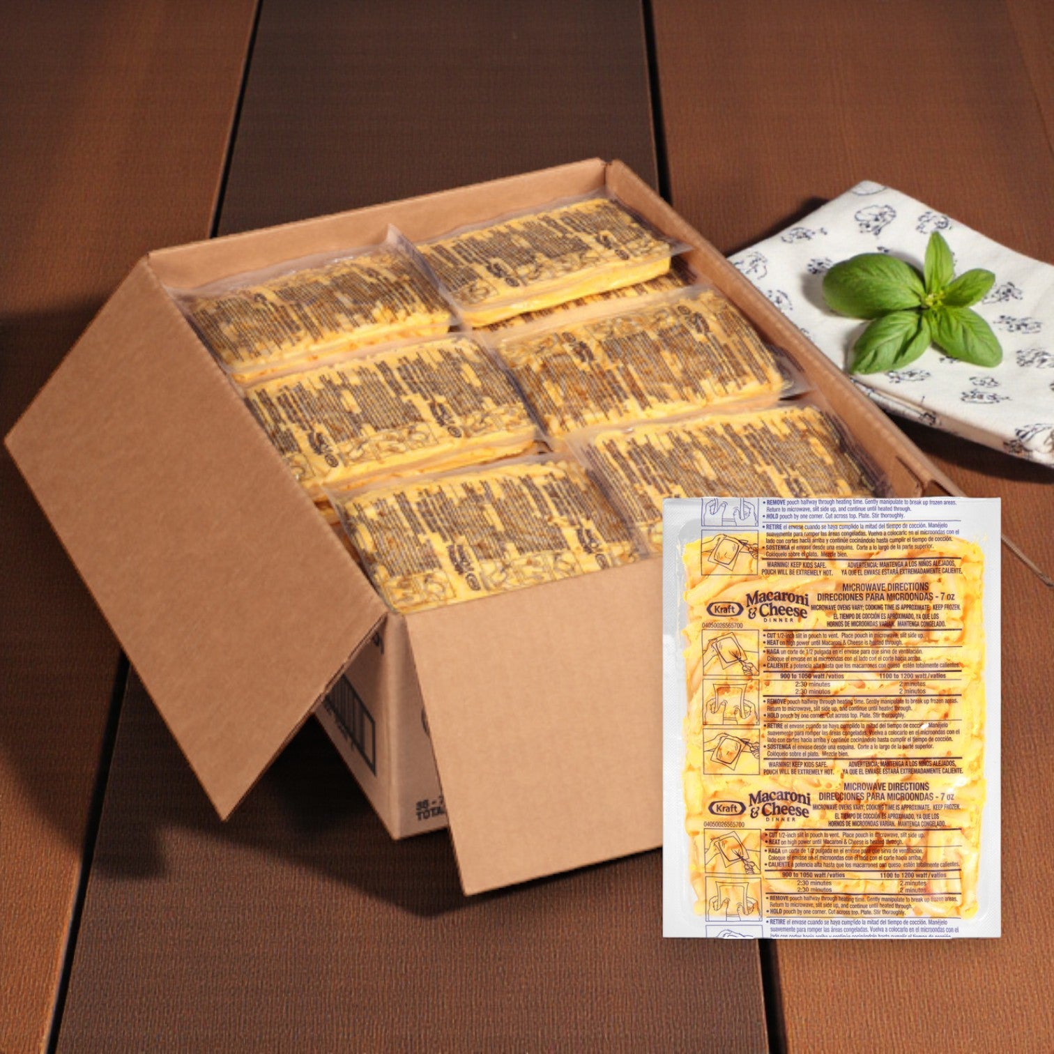 An open cardboard box containing Kraft Frozen Entree Macaroni & Cheese – 36 Packs (7 oz each) sits on a wooden table beside a folded napkin and a sprig of basil.