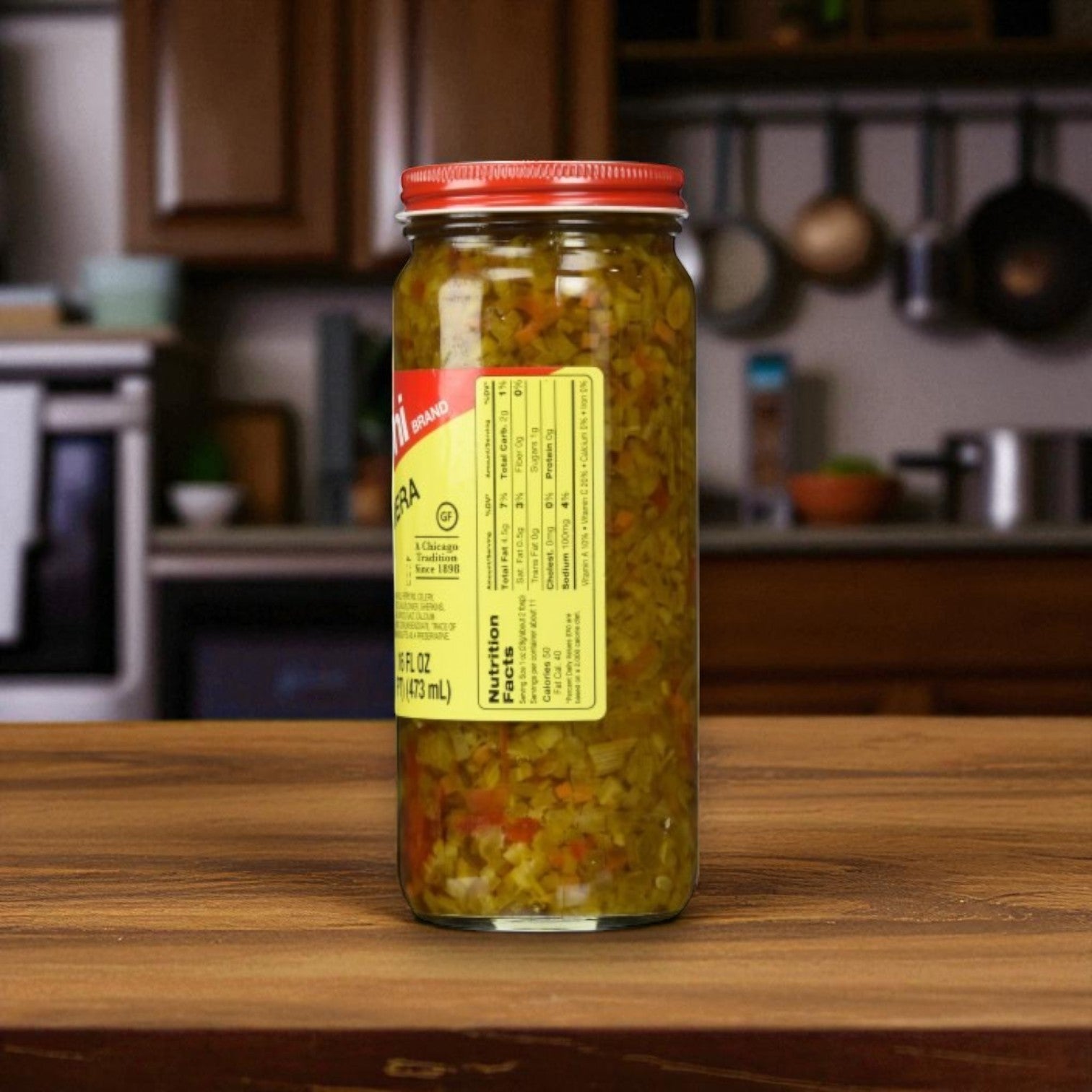 A 16 oz jar of Marconi Brand Giardiniera – Mild from Macroni Foods, with a red lid, sits on a wooden counter showing its yellow nutrition label. Kitchen appliances and utensils are visible in the background.