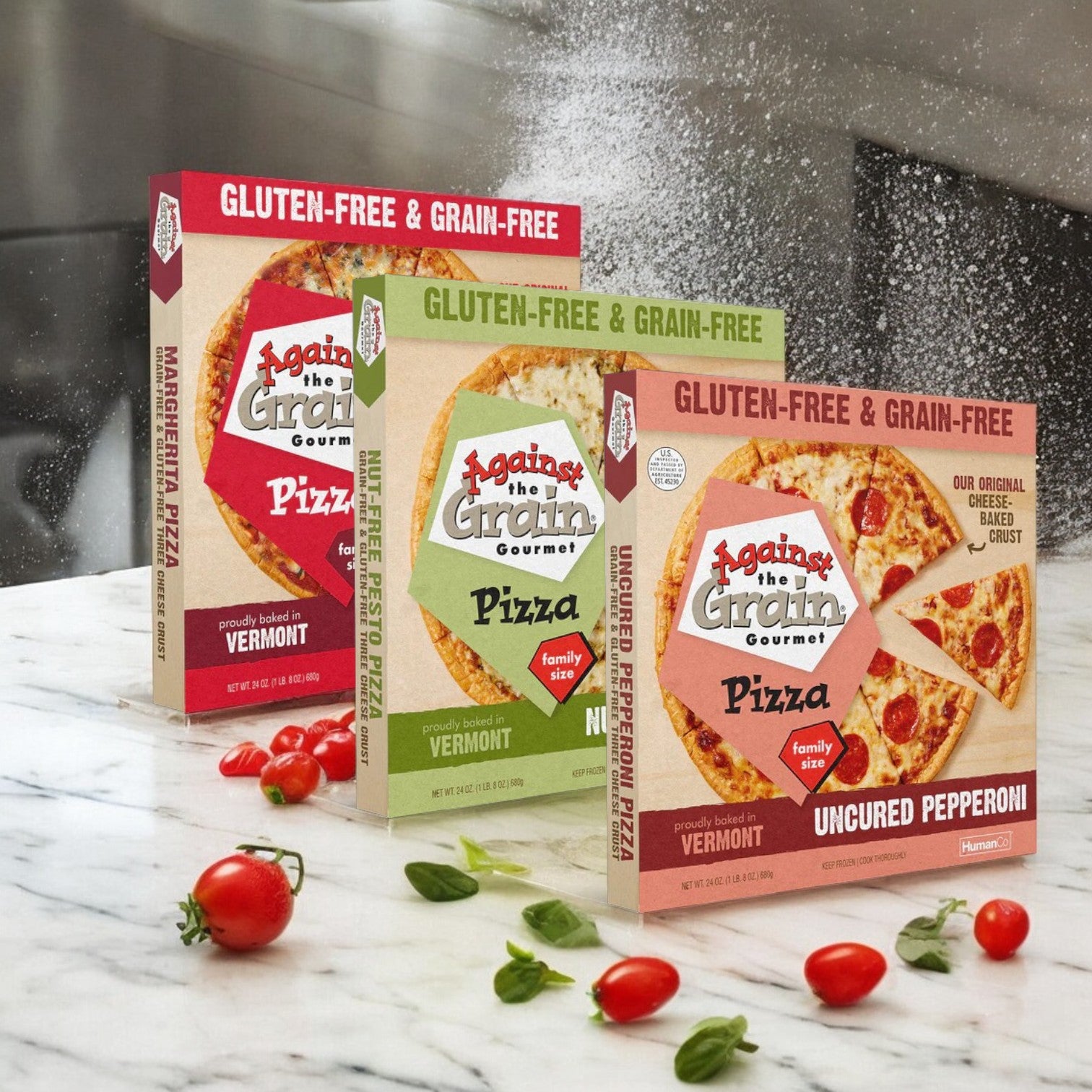 A 3-Pack Combo of Against the Grain Gourmet Pizza is displayed on a marble surface, surrounded by small tomatoes and a hint of flour dust in the air.