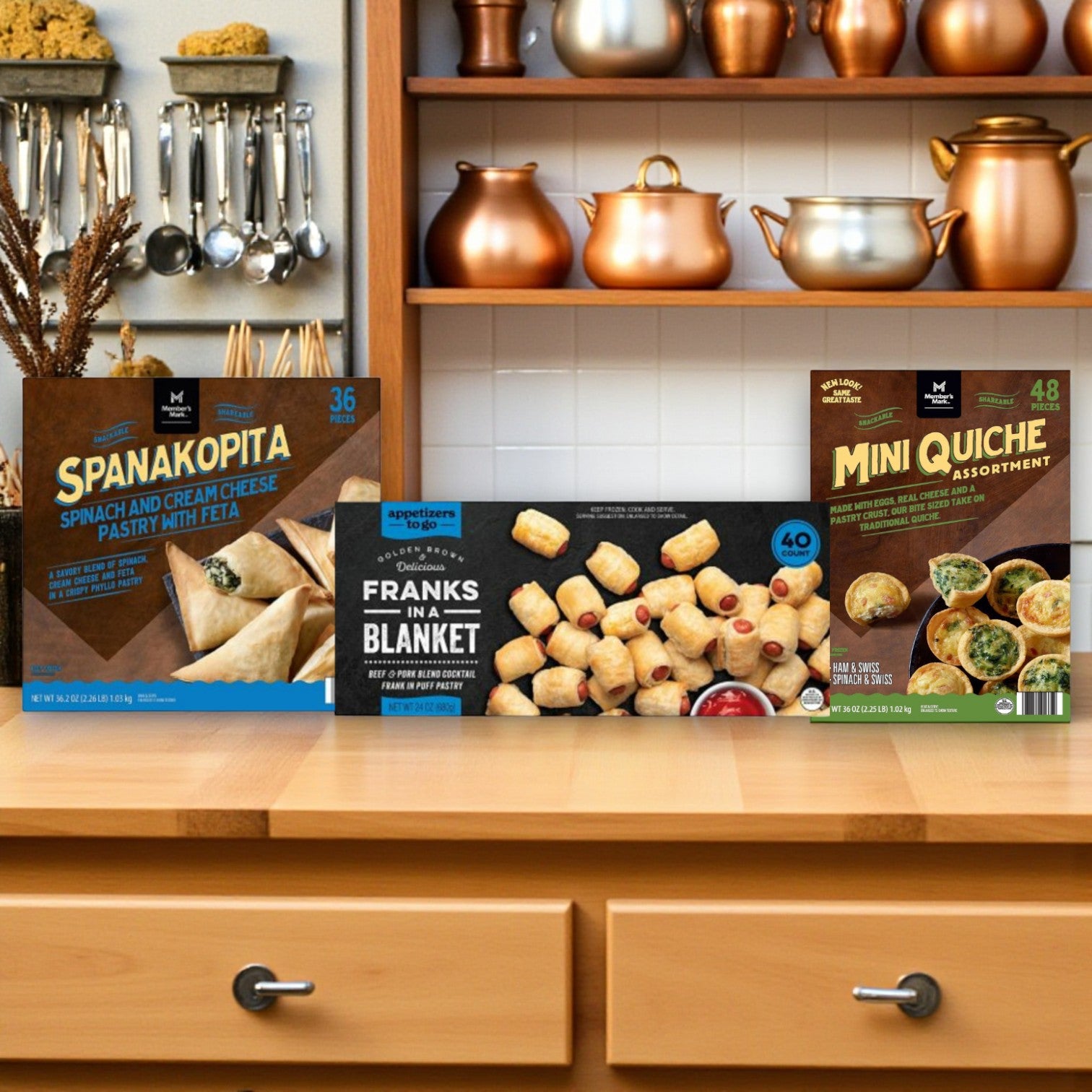 Three boxes of Easy Lunches Member’s Mark Appetizer Party Combo—Mini Quiche, Spanakopita, and Franks in a Blanket—are arranged on a wooden kitchen counter with shelves and copper cookware in the background.