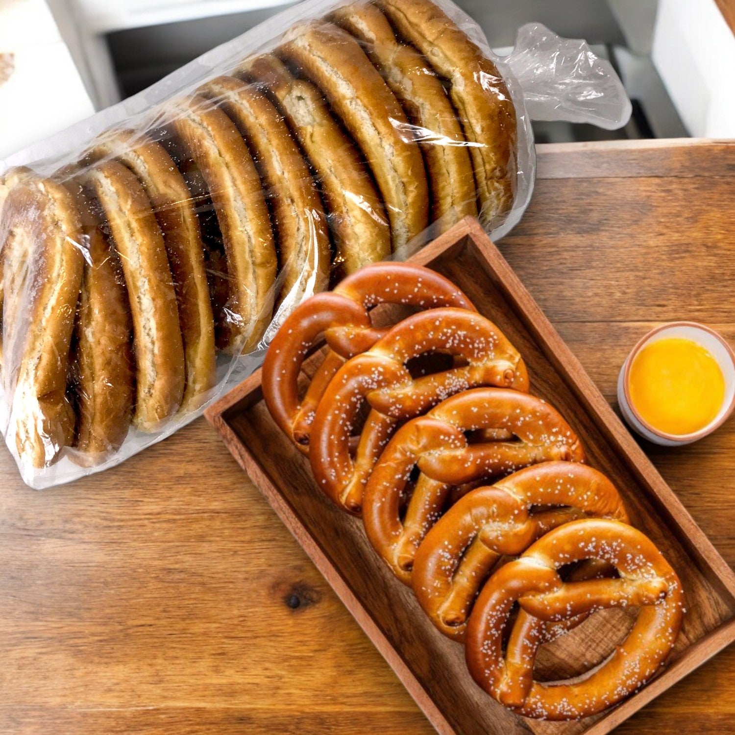 Milwaukee Pretzel Company Bavarian Twist Pretzels sit alongside flavorful bagels on a wooden tray with a small cup of creamy cheese dip on a rustic table.