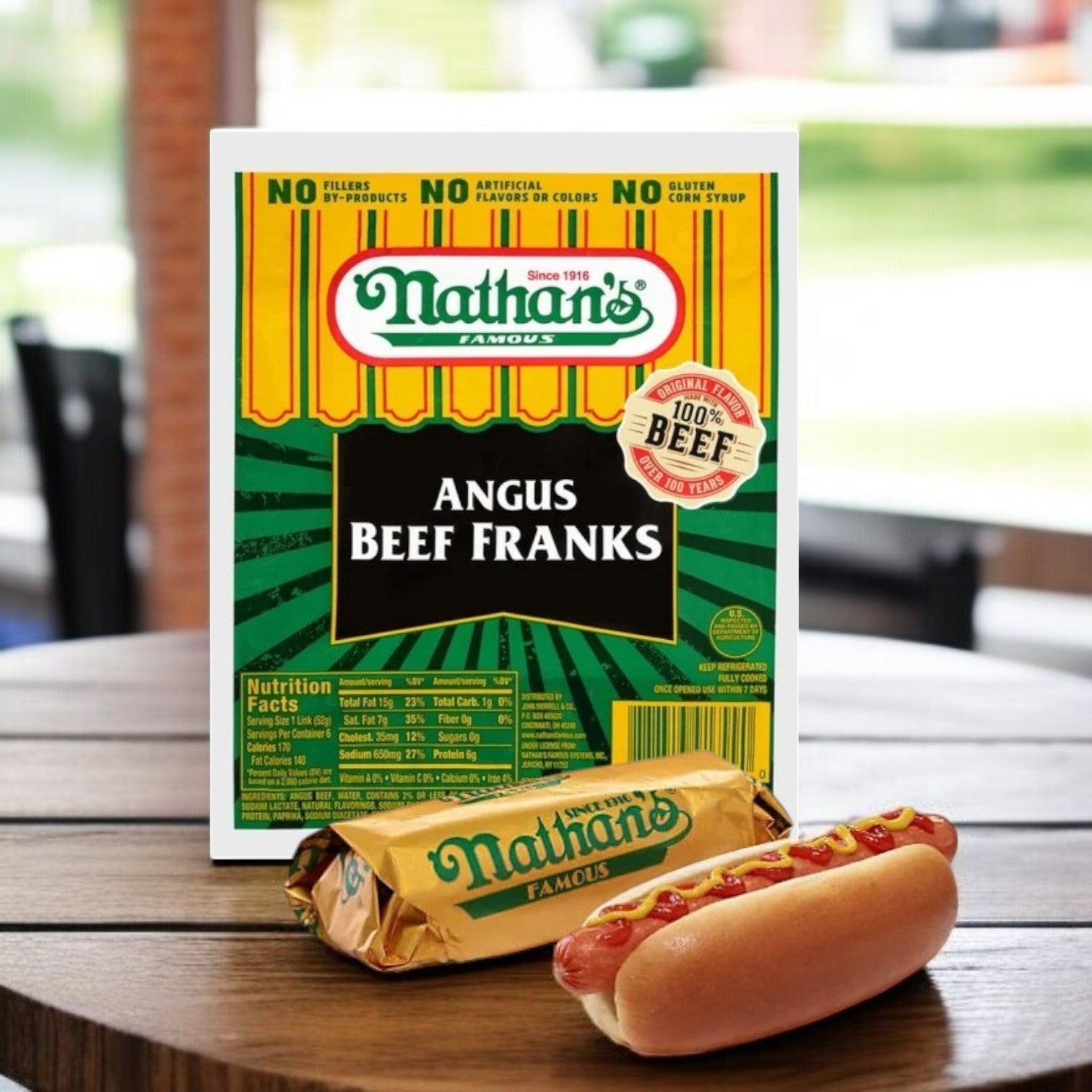 A Nathan’s Famous Angus Beef Franks – 11 oz Pack (6 Hot Dogs) box sits behind a wrapped and an unwrapped all-beef hot dog in a bun on the table.