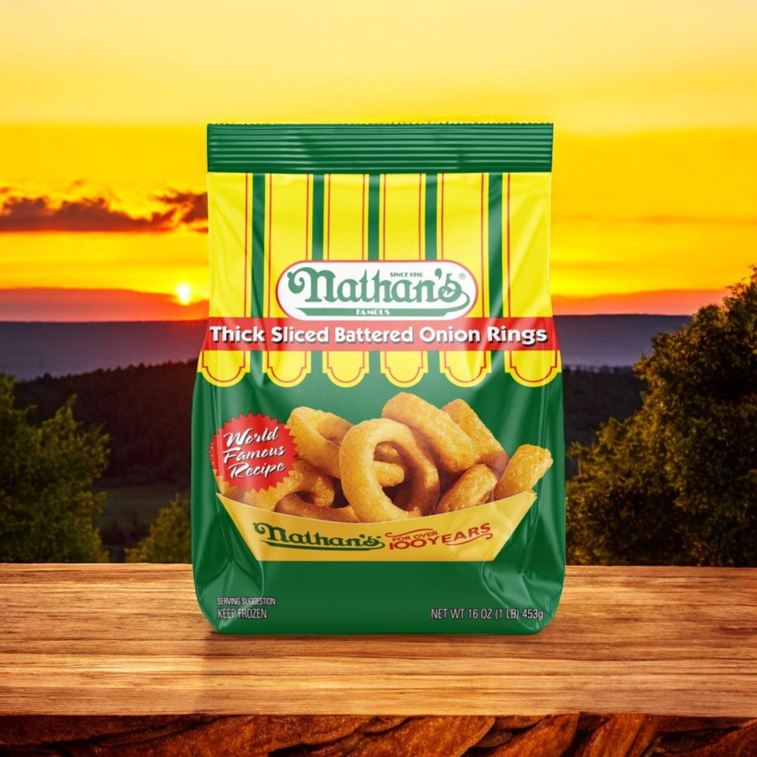 Three 16 oz bags of Nathan’s Famous Thick-Sliced Battered Onion Rings sit outdoors on a wooden surface, highlighting the crispy, thick onion rings with a scenic sunset and trees in the background.