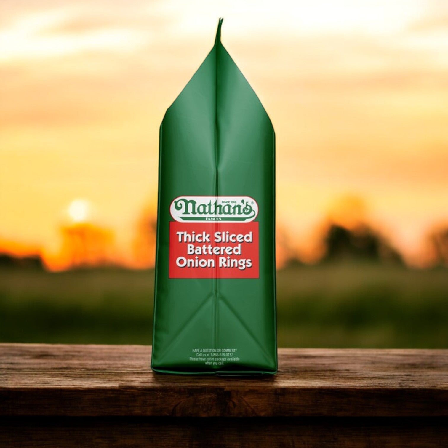 Three 16-oz bags of Nathan’s Famous Thick-Sliced Battered Onion Rings are displayed on a wooden surface, their green packaging highlighting crispy, thick-cut rings with a sunset in the background.